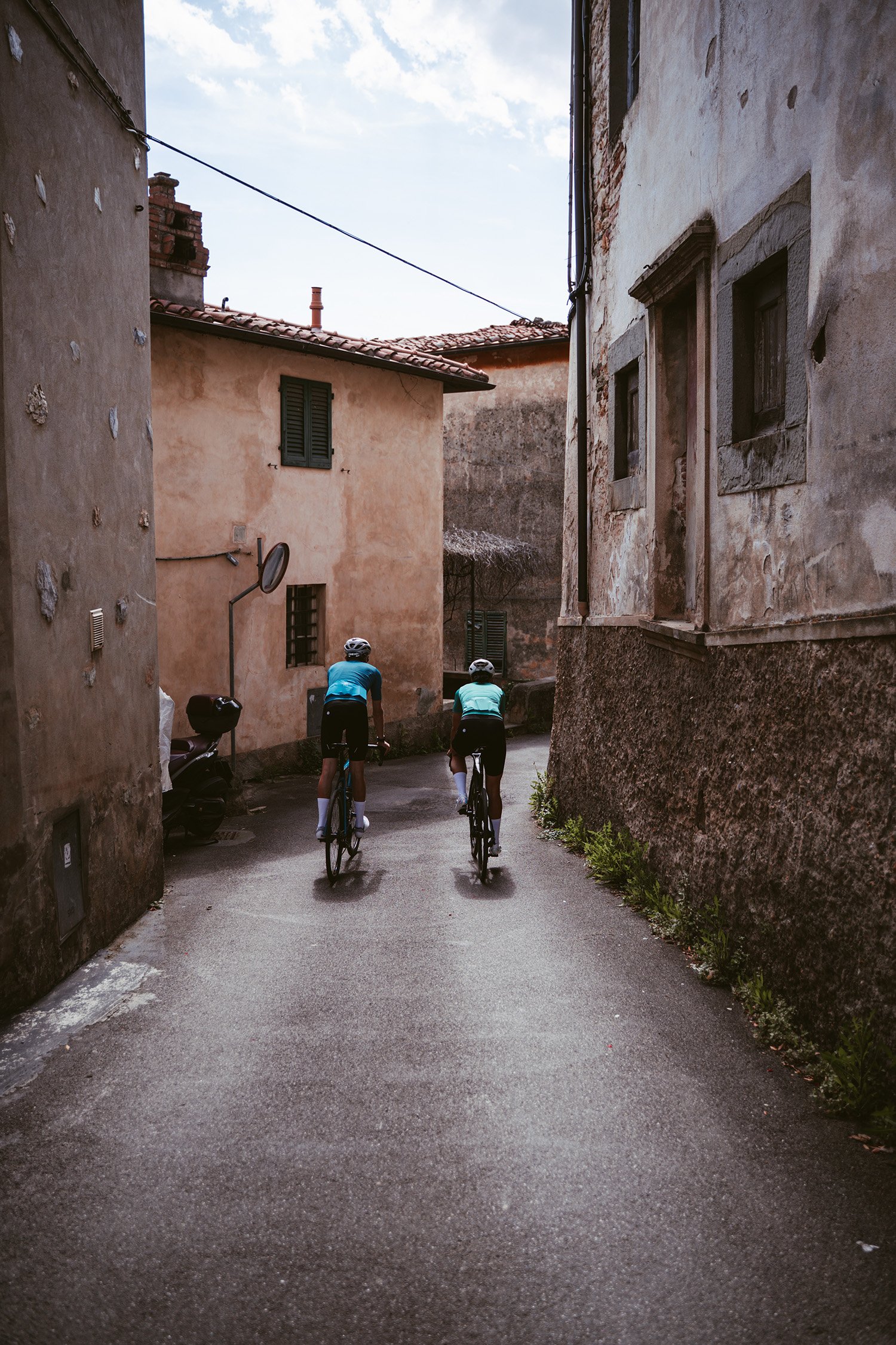 Two cyclists riding uphill through a narrow, rustic alleyway flanked by weathered buildings with peeling paint and small windows, under a partly cloudy sky.