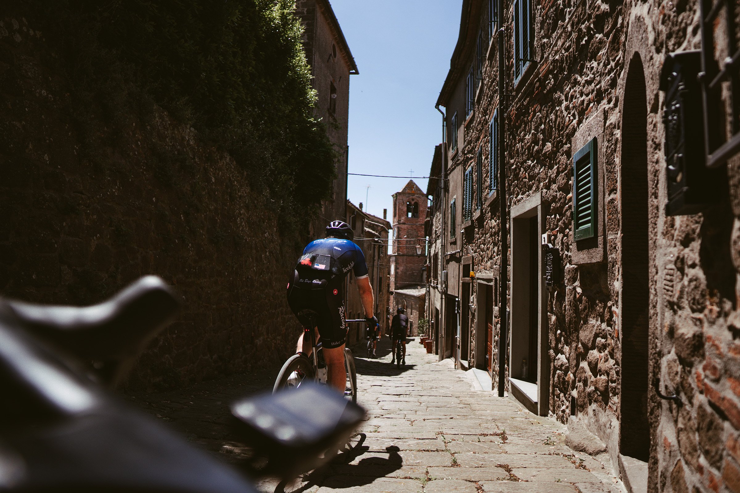 A narrow cobblestone street in a historic European town with brick and stone buildings on either side, and a cyclist riding away from the camera towards a bell tower in the background under a clear blue sky.