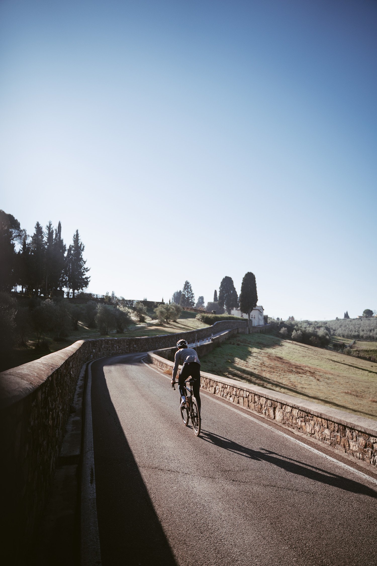 A cyclist riding up a winding, shadowed road in a hilly, rural landscape under a clear blue sky.