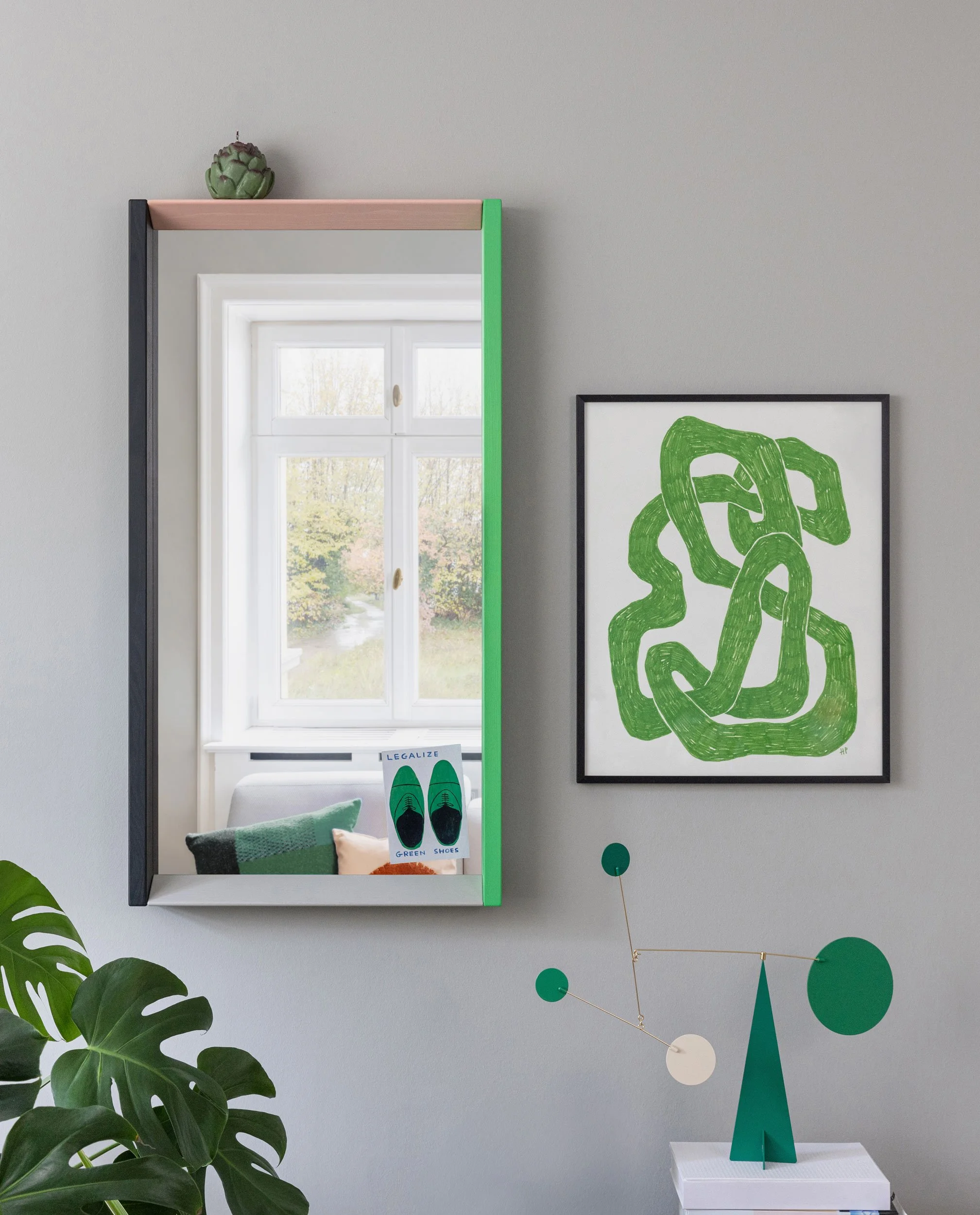 Color Frame Mirror. Spiegel  in grün/rosa an Wand. Davor ist eine Zimmerpflanze und ein Mobilé in grün