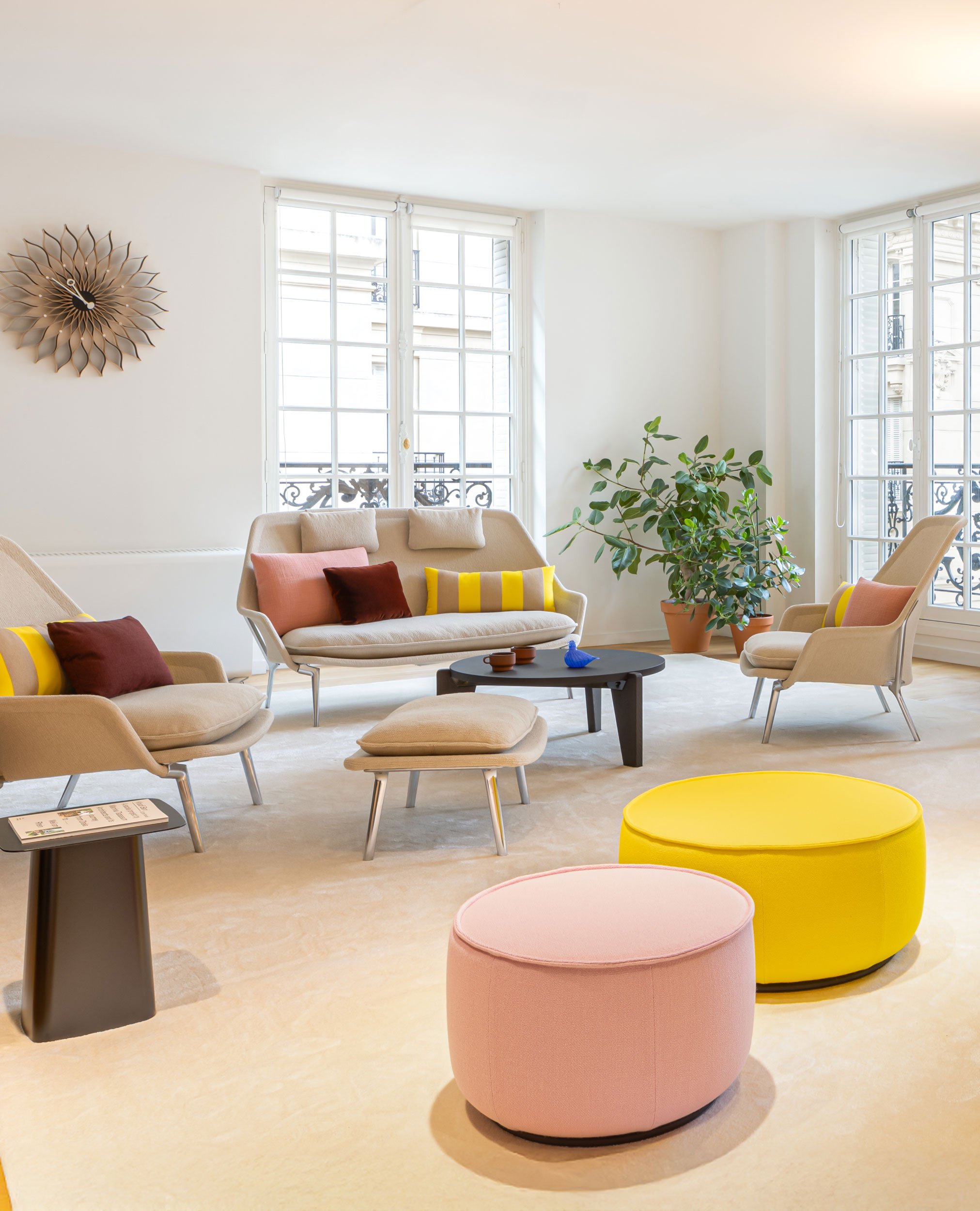 Slow Sofa und Slow Chair Landschaft mit farbigen Kissen und gelbem und rosa Mariposa Ottoman. Creme farbiger Teppich. Pflanze in Ecke und Sunflower Clock an Wand