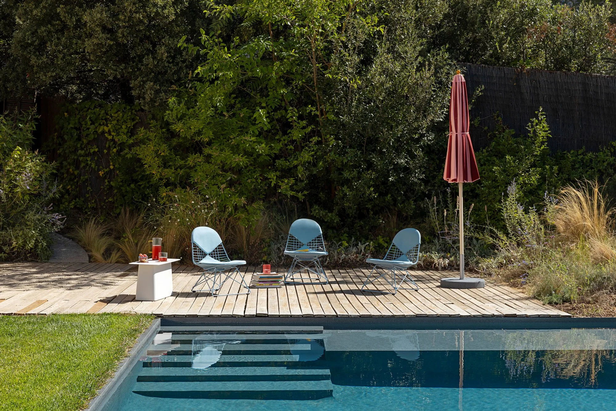 Drei hellblaue Wire Chairs stehen am Pool. Ein roter Sonnenschirm steht daneben.
