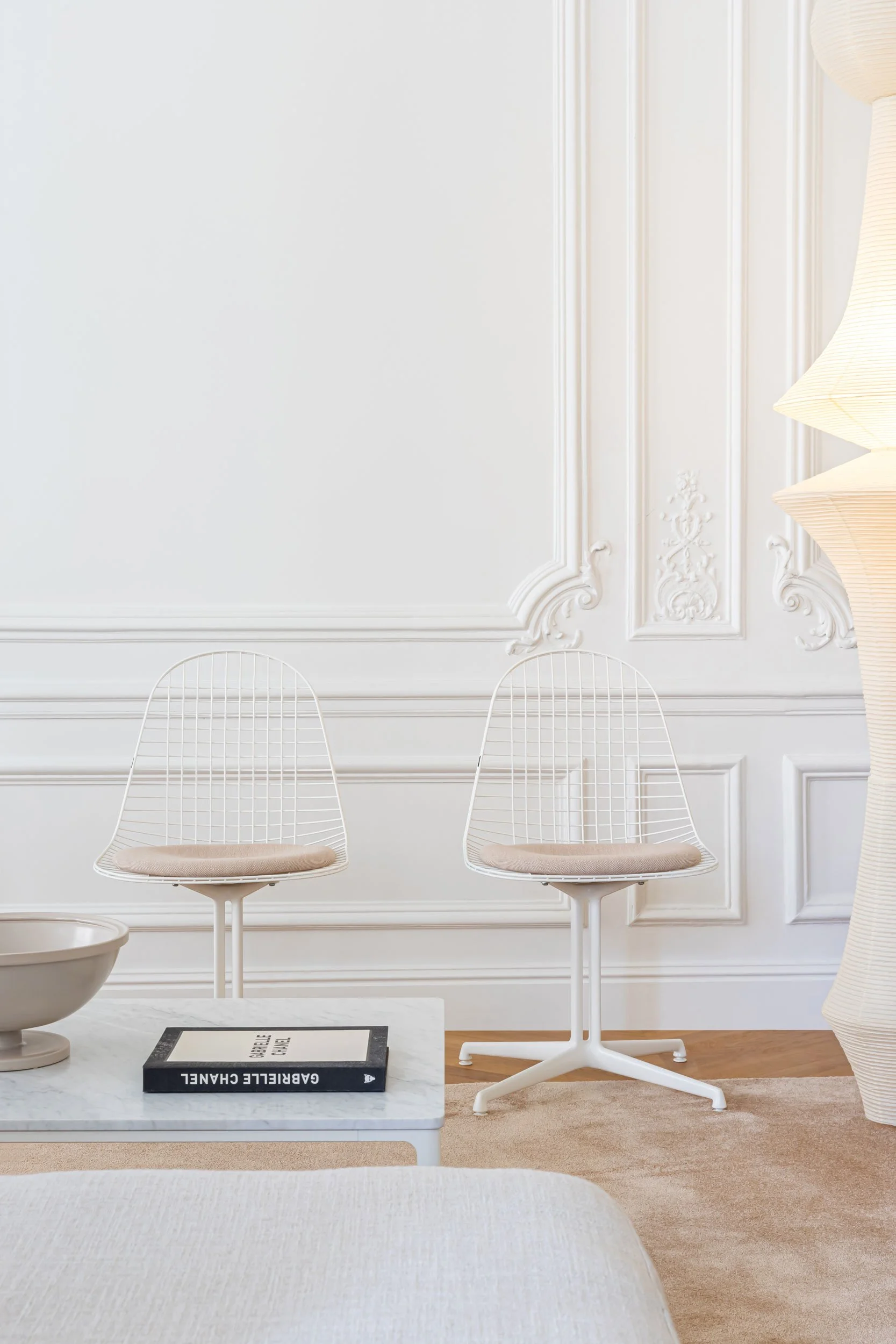 zwei weisse Wire Chairs mit La Fonda Fuss vor weisser Wand mit Stuckaturen, Akari angeschnitten an Seite, Plate Table mit Buch und Schale davor. Teppich in creme