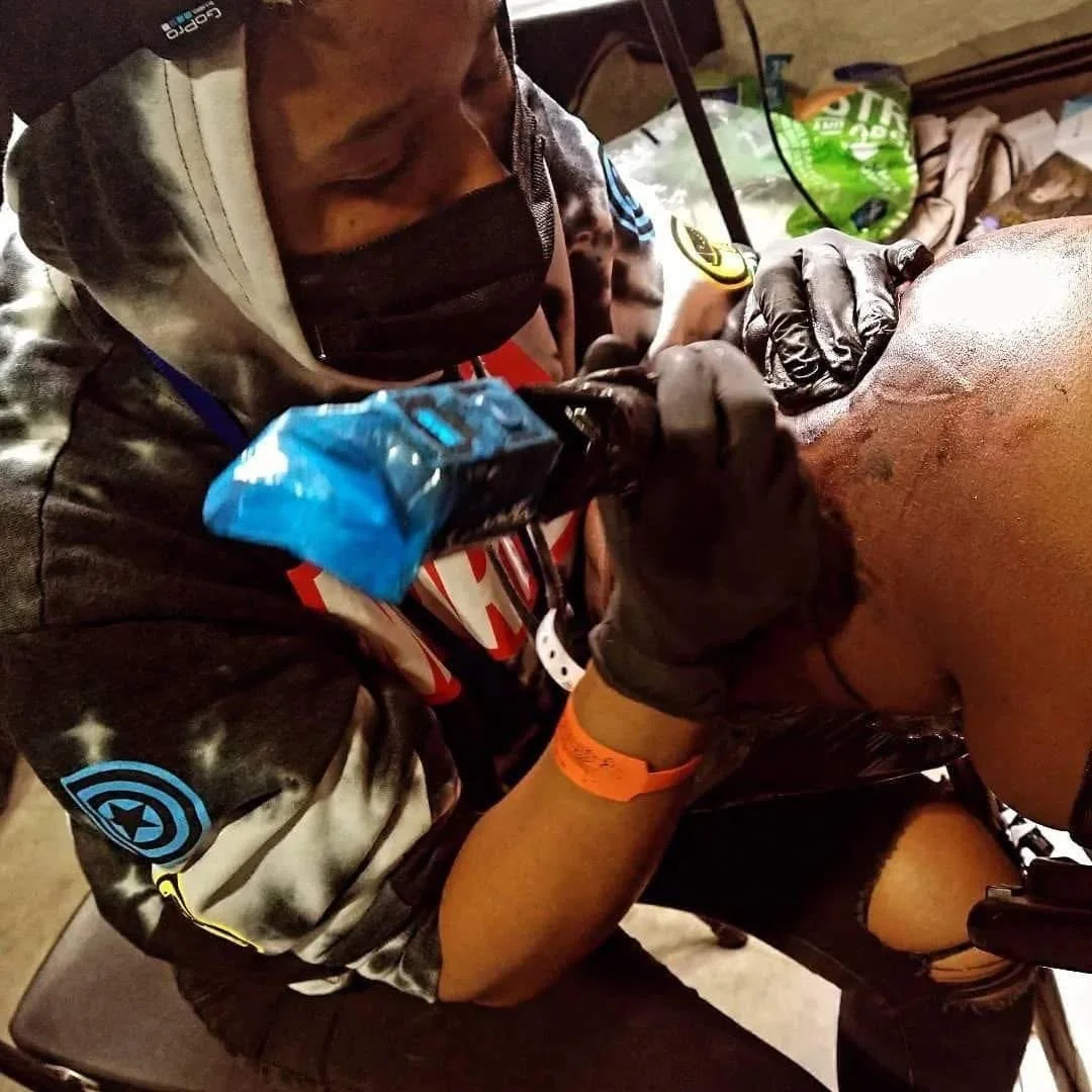 Tattoo artist working on a person's arm in a tattoo shop, with supplies and tools visible in the background.