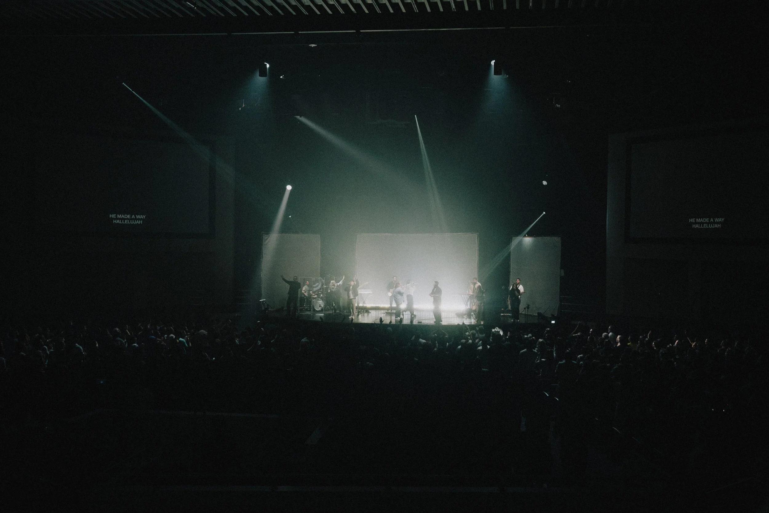 A dimly lit worship concert scene with a band on stage and a large live audience in the foreground, surrounded by dramatic lighting effects.