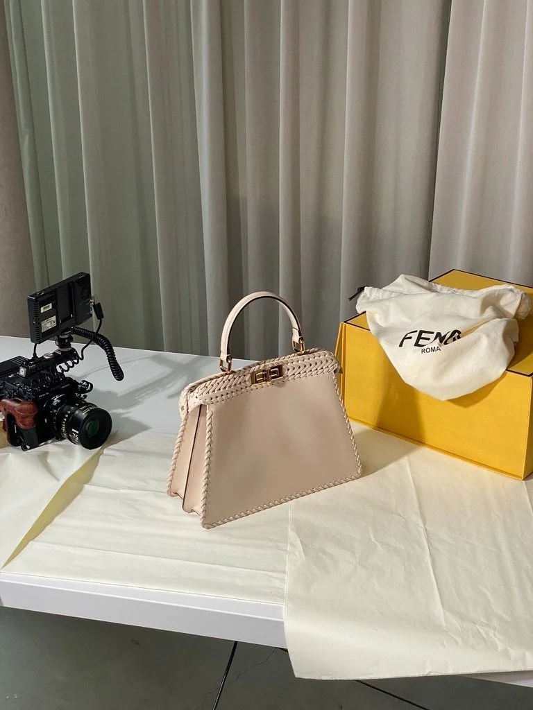 A professional camera, the Fendi Roma Peekaboo handbag, yellow box and dust bag sits on top of a table with a curtain hanging behind it.