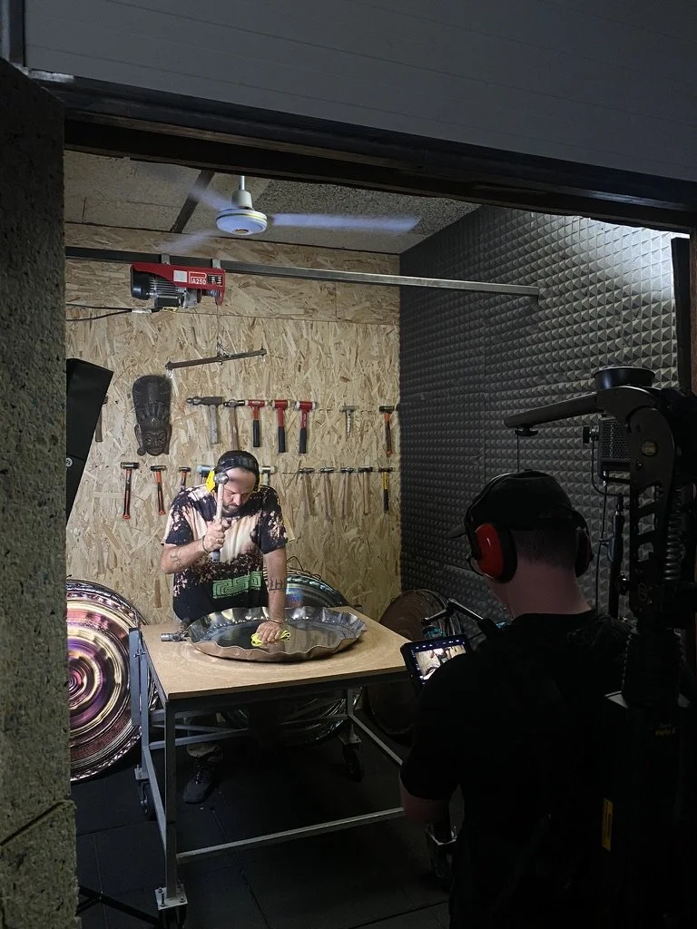 Behind-the-scenes of videographer with professional camera and headphones capturing gong-maker hammering metal plate in a workshop with bright lighting and tools mounted on the wall behind him.