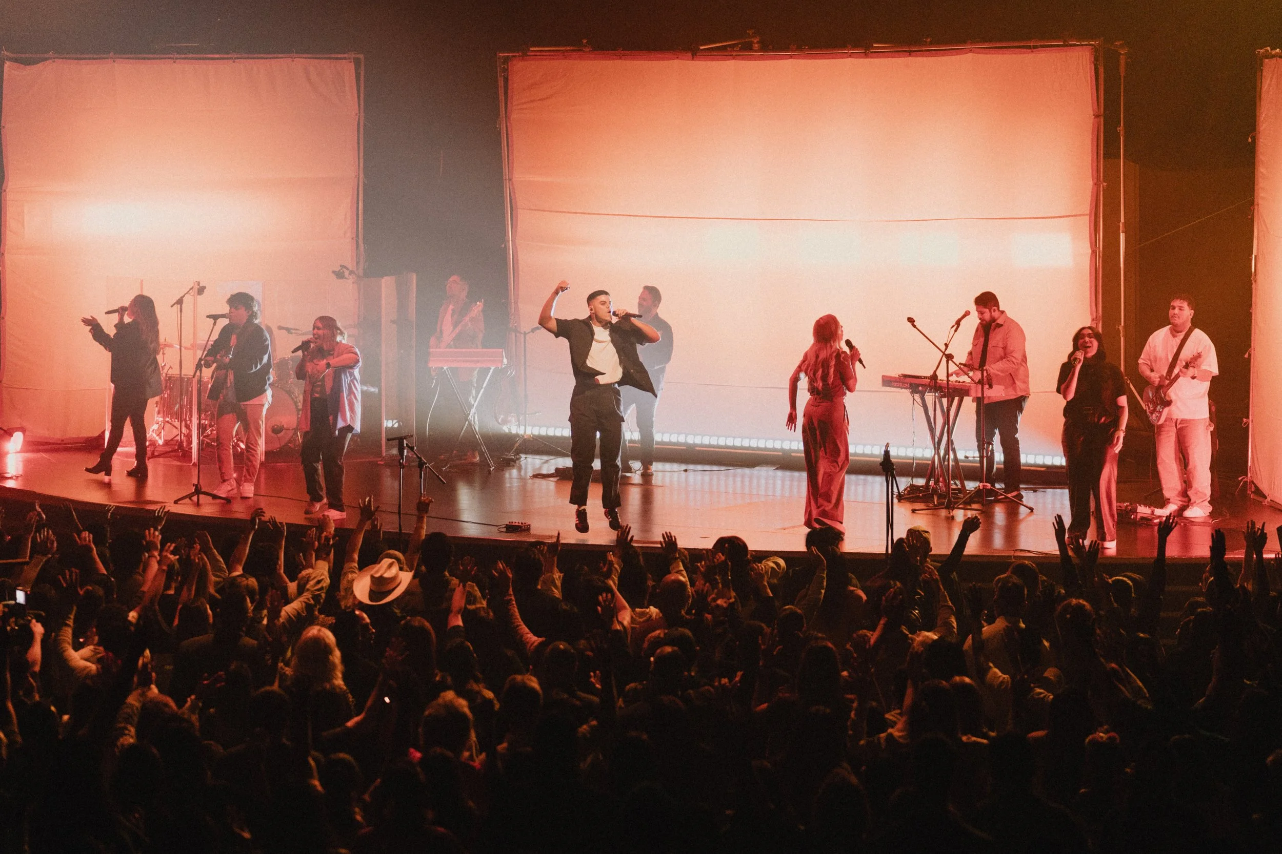 A worship team on stage, surrounded by a singing live audience, and illuminated by bright lighting with screens behind them. 