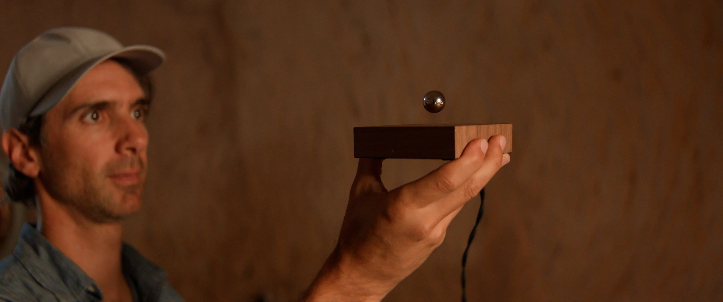 A man wearing a gray cap holds a wooden block with a metal ball above it illustrating acoustic levitation using sound waves, against a brown stone background.