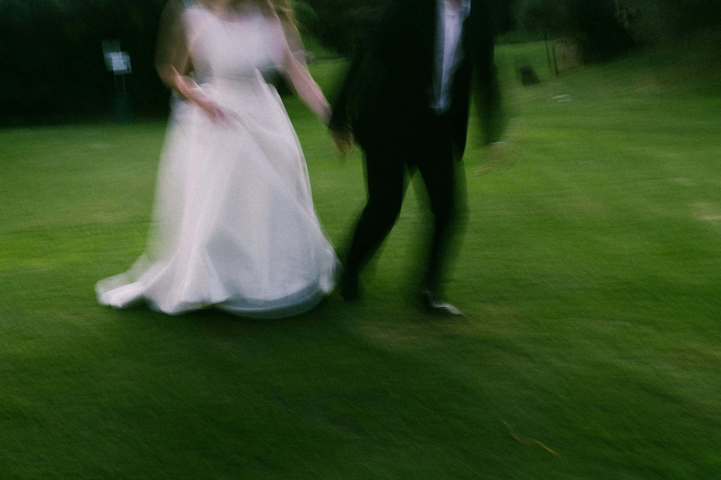 A blurred image captured on film of a bride in a white wedding dress and a groom in a dark suit holding hands while running on grass.