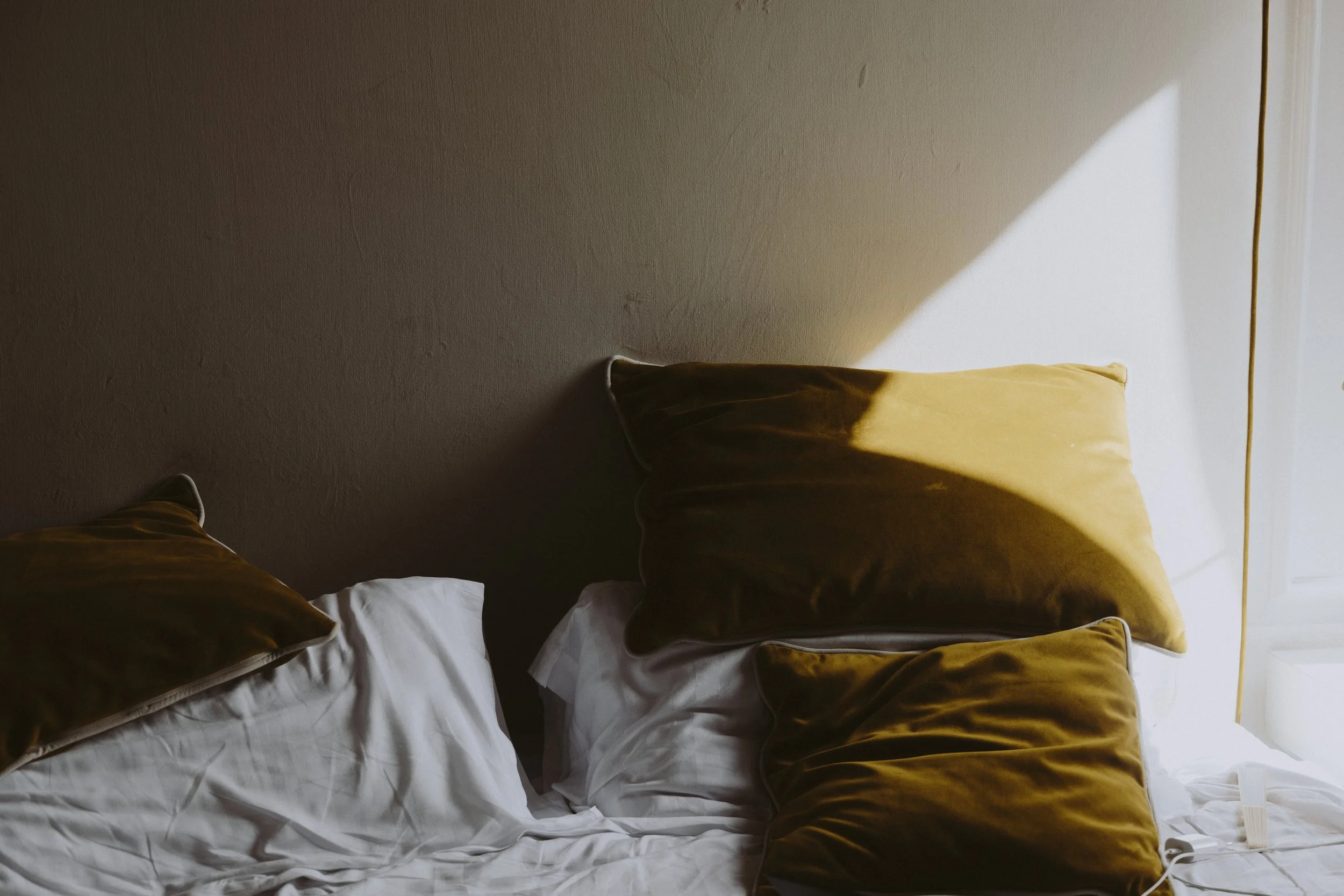 A bed with white sheets and mustard-coloured pillows and scatter cushions against a wall, with soft sunlight breaking through the window, illuminating part of the scene.