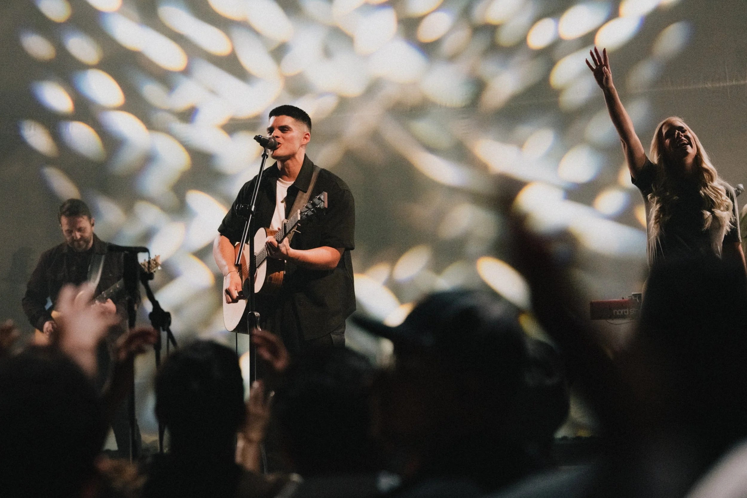 Worship team singing on stage in front of a live crowd with white textured visuals behind them.