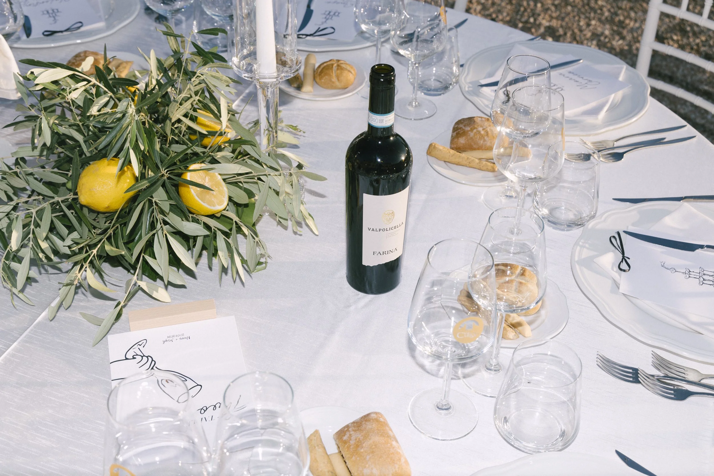A beautifully arranged wedding dining table setting with glasses, bread rolls, a bottle of wine, and a centerpiece of lemons and greenery.