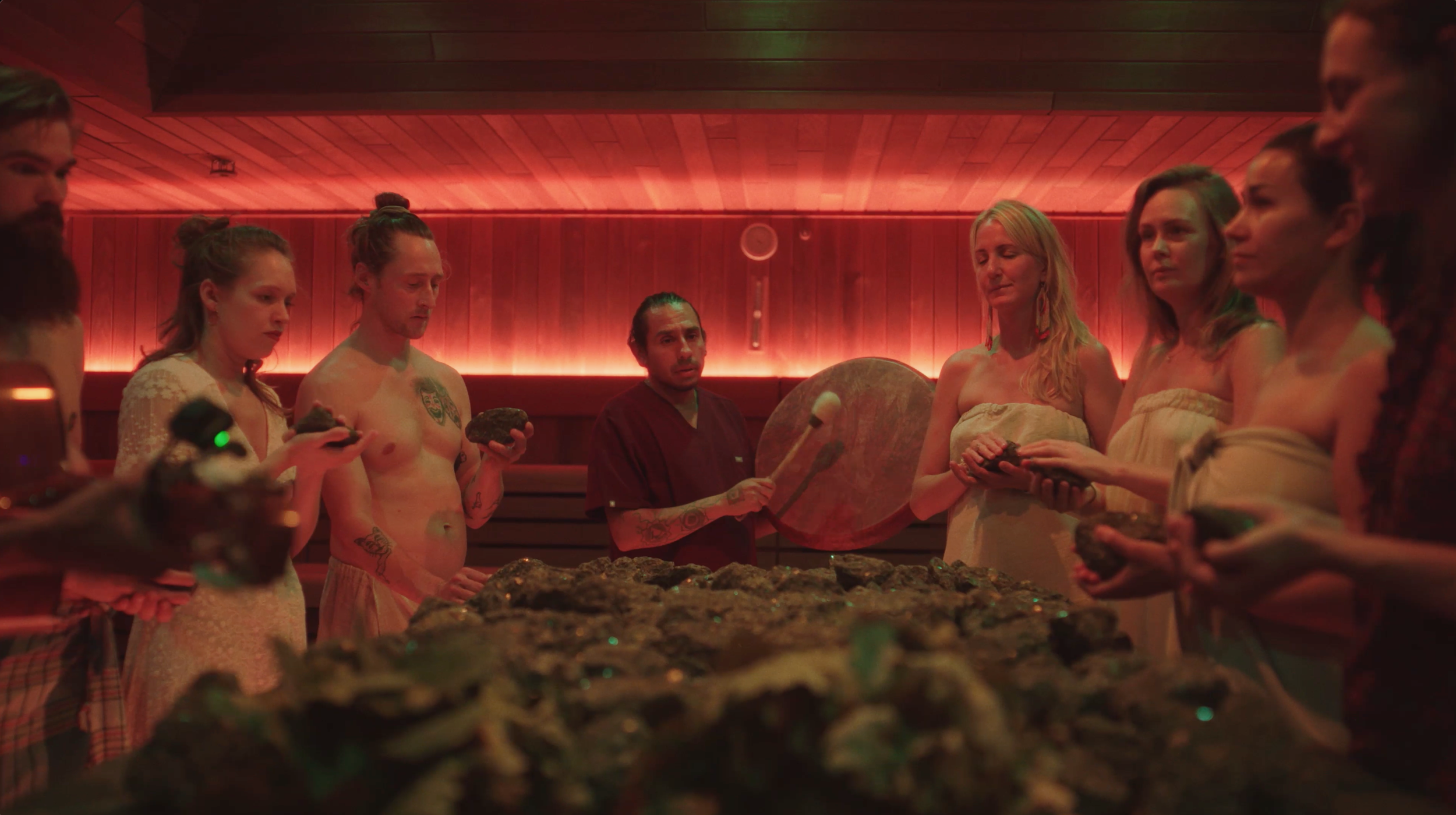 A group of people stand in a dimly lit room with red lighting, holding rocks and a gong, gathered around a central table covered with rocks as part of a spa ritual.