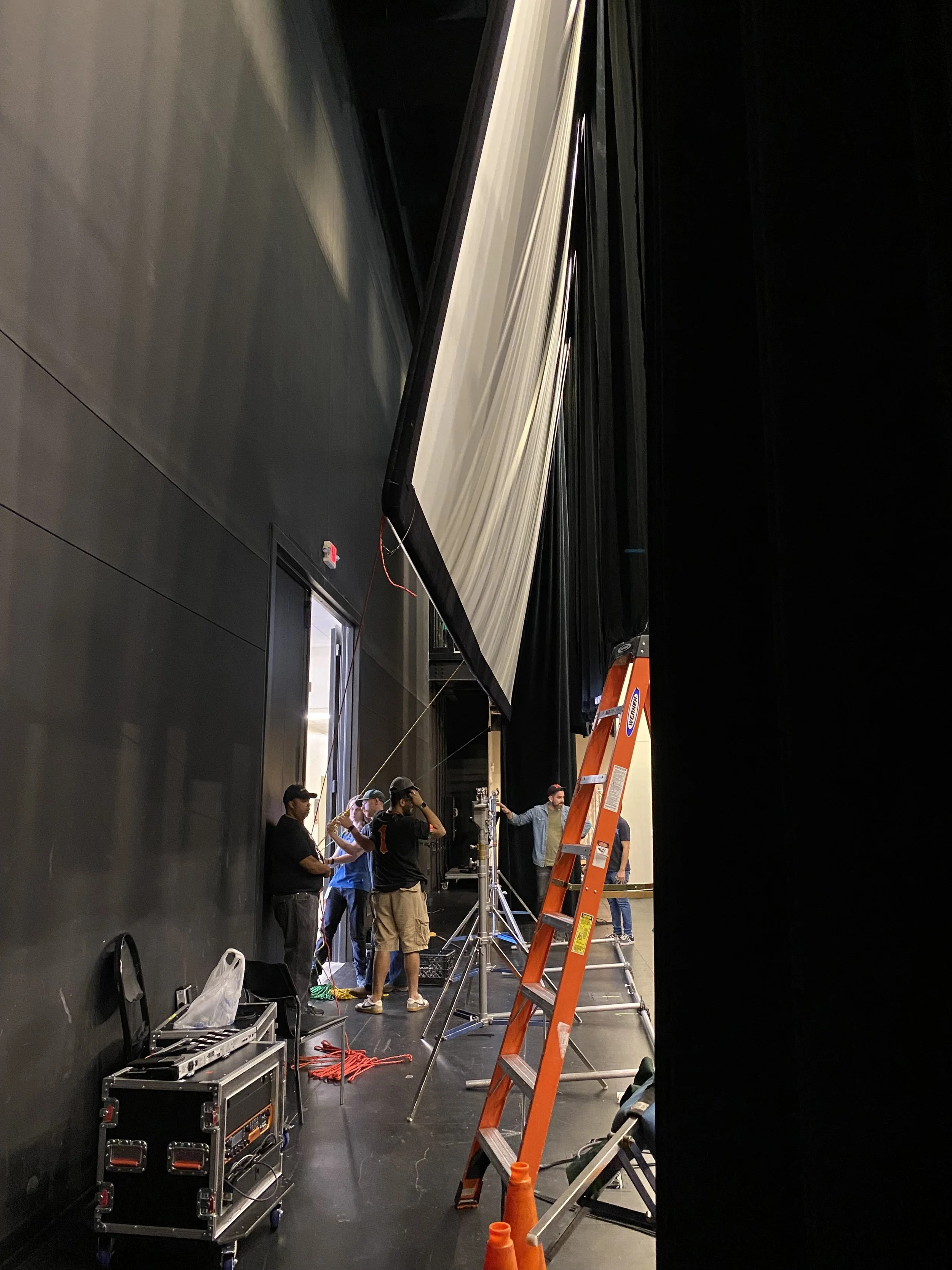A group of people setting up equipment on a stage, with a tall ladder and a large screen being installed.
