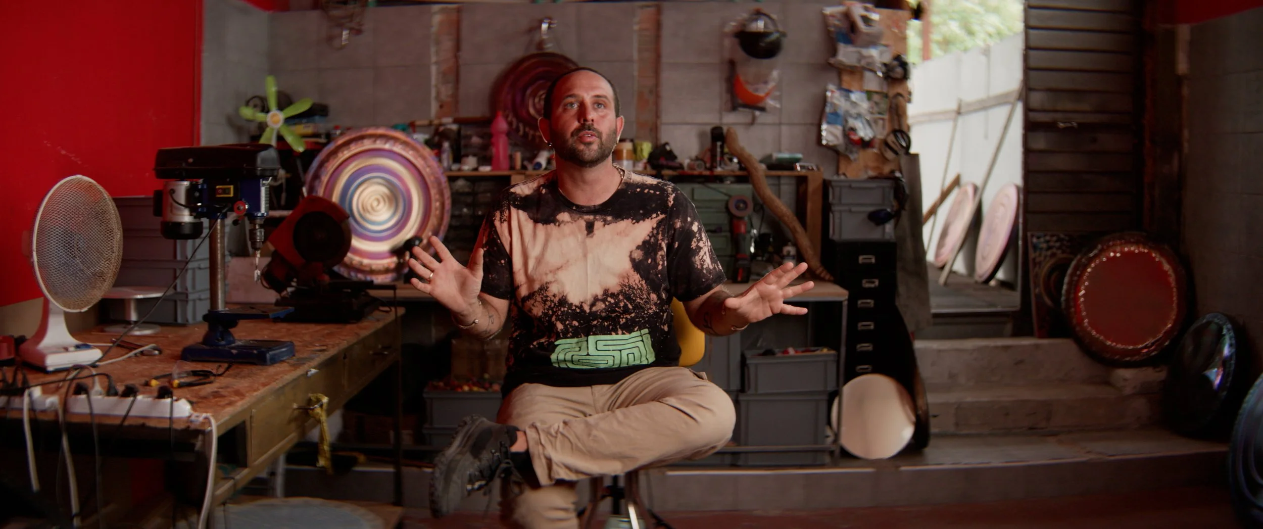 A man, wearing a black and beige tie-dye t-shirt and beige pants, sitting cross-legged on a stool in a workshop space during an on camera interview with various artwork, tools, gongs and materials in the background with a red wall to his right and a