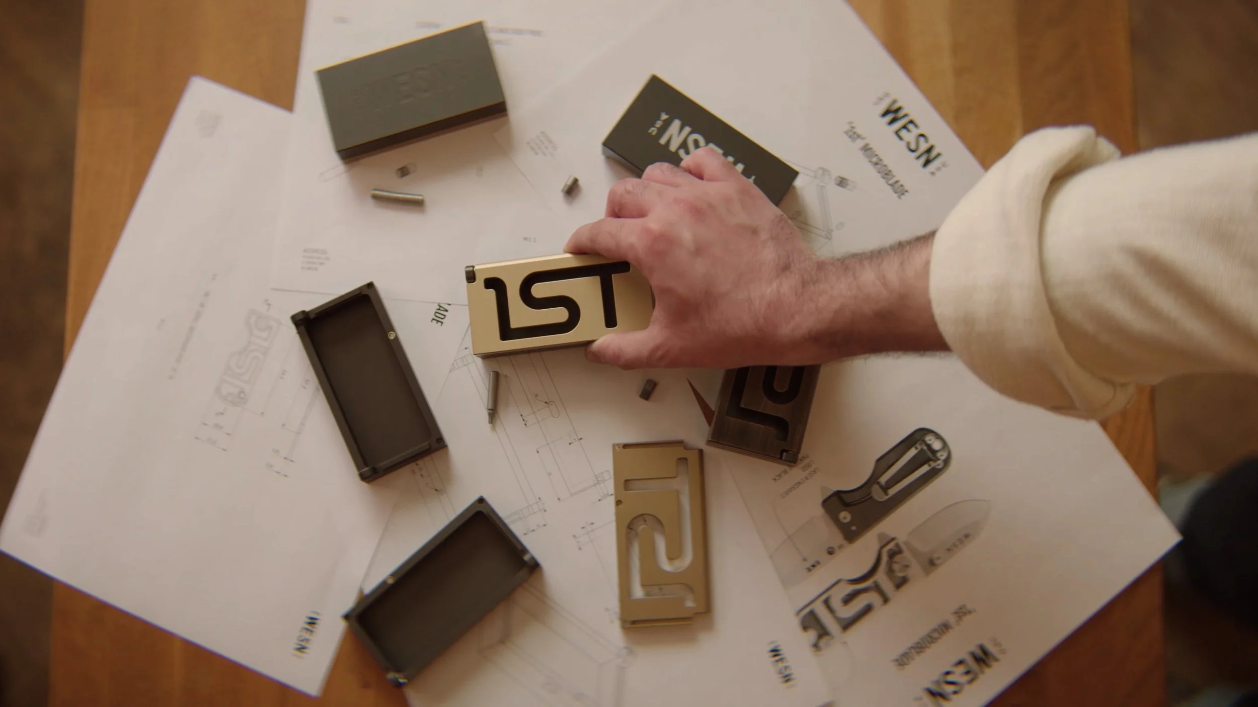 A person's hand reaches for a small, metallic hilt labeled "1st" on a wooden table scattered with design blueprints and pocket knife crafting materials part of the planning and design process.