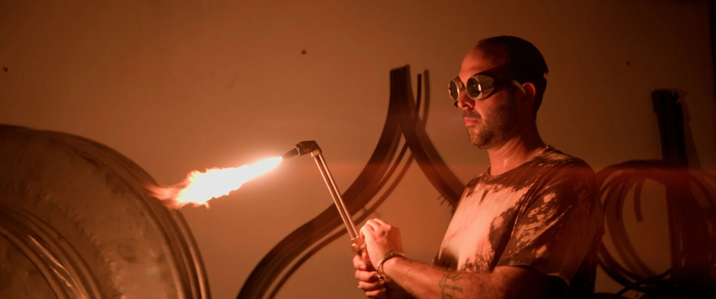 Man wearing safety goggles holds a flame torch in the air as part of gong-making process with gongs and tools in the background mounted on a stone wall.