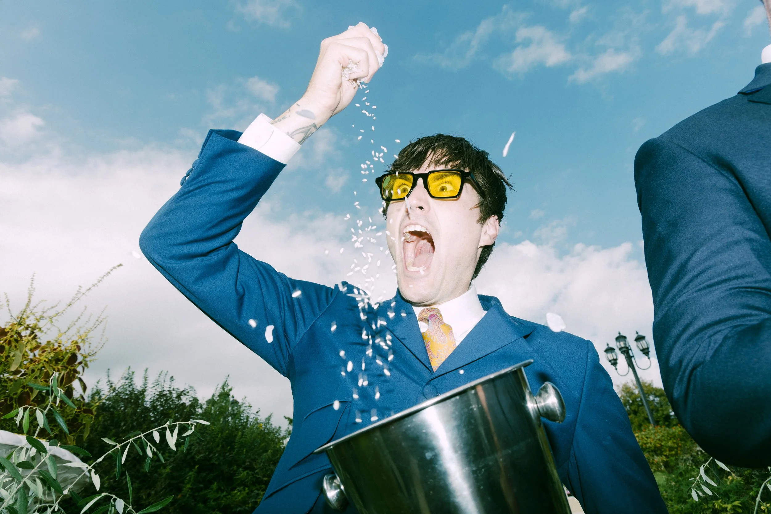 A man in a blue suit and yellow sunglasses is excitedly throwing ice into a metallic ice bucket, with an expressive facial gesture, against a cloudy sky backdrop with trees. 