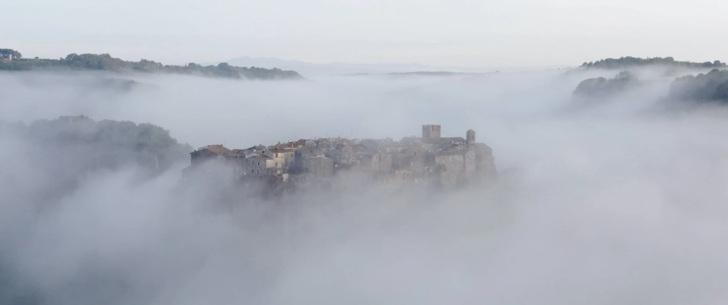 A foggy landscape with a historic stone castle on a hilltop surrounded by mist, clouds and distant rolling hills.