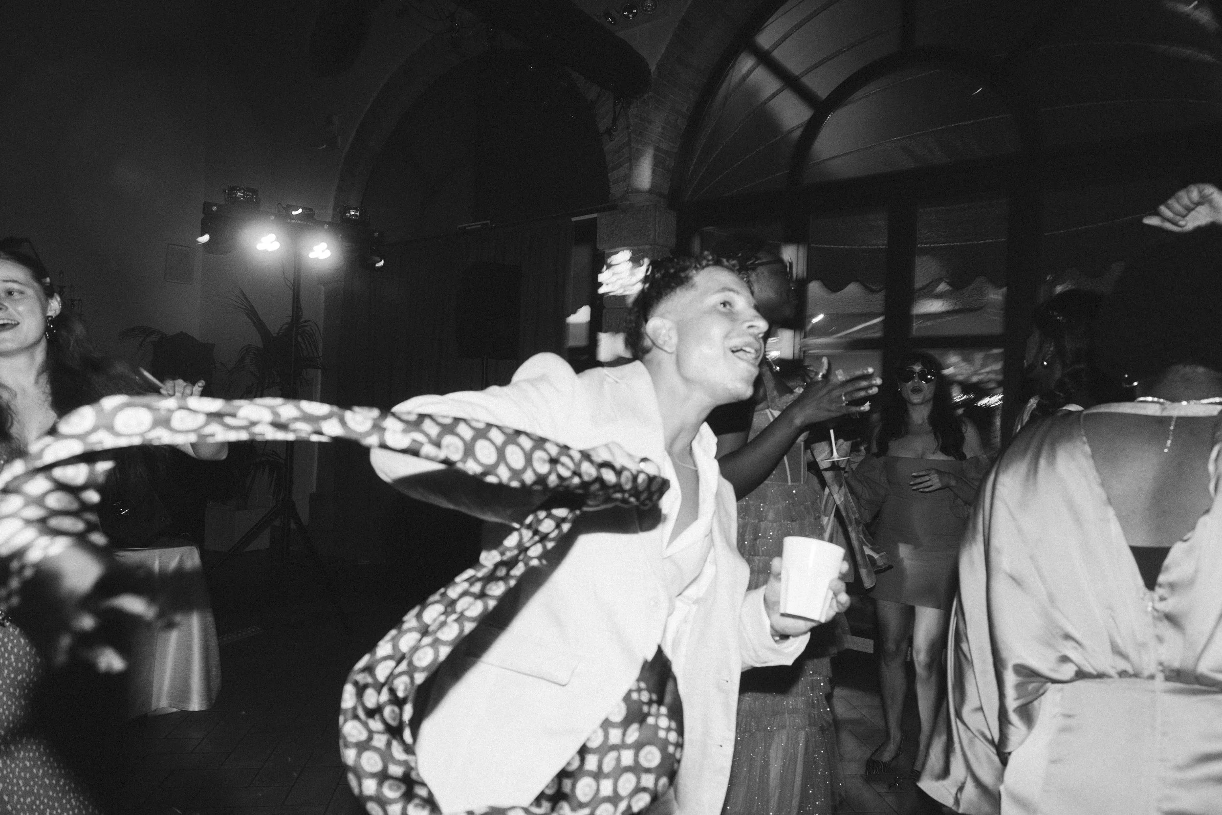 A black and white photo of a lively party scene where people are dancing. One person in the foreground is wearing a light-colored suit and holding a cup, while other guests can be seen in the background enjoying the wedding festivities.