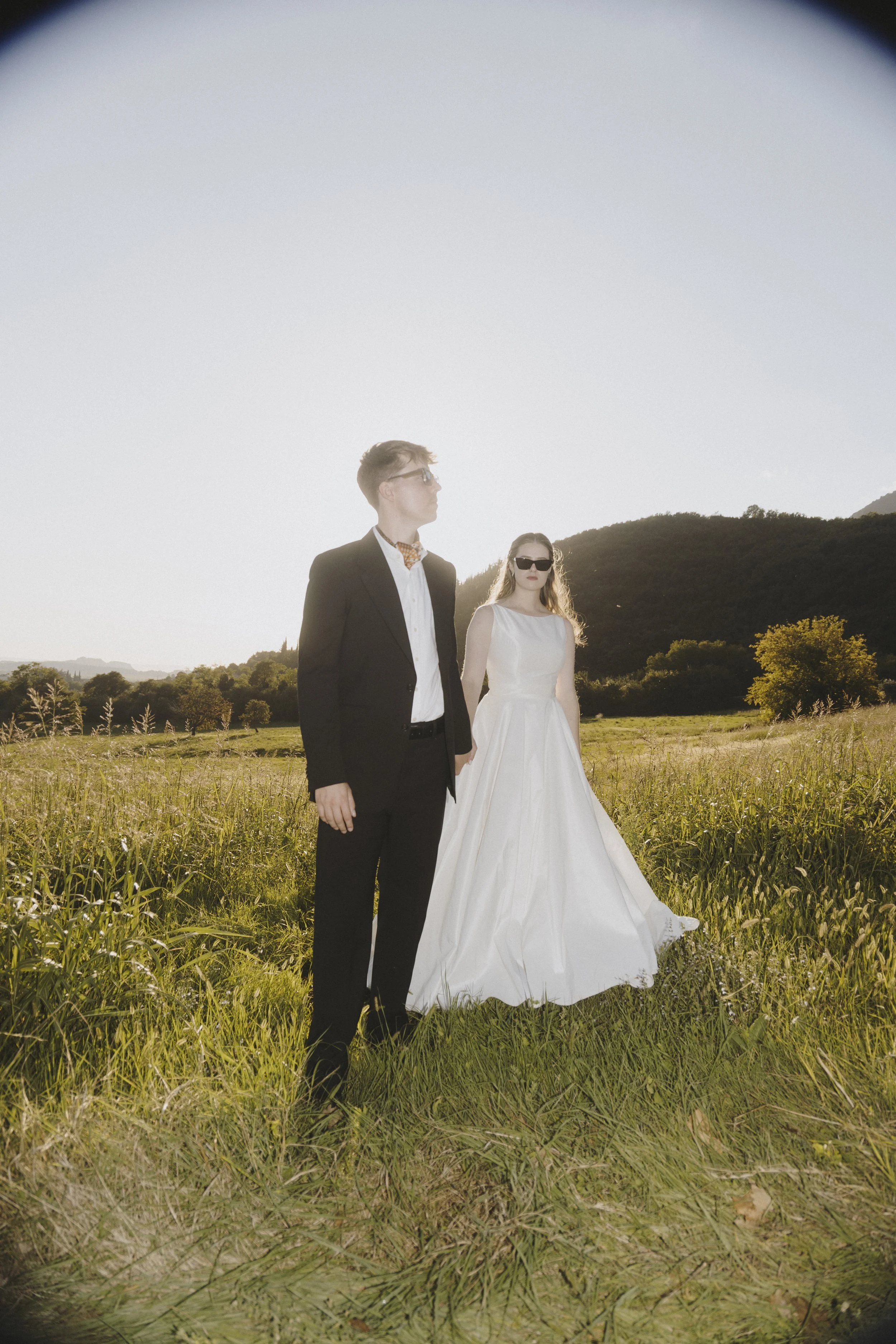 A groom and bride standing in a grassy field, the sun shining brightly behind them. The man is dressed in a black suit, while the woman wears a white wedding dress and sunglasses. Mountains are visible in the background. 