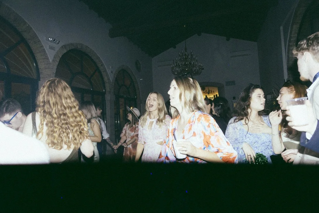 A lively wedding scene with people dancing and socializing, featuring a mix of outfits and hairstyles in a historic building with arched windows in the background, captured on film.