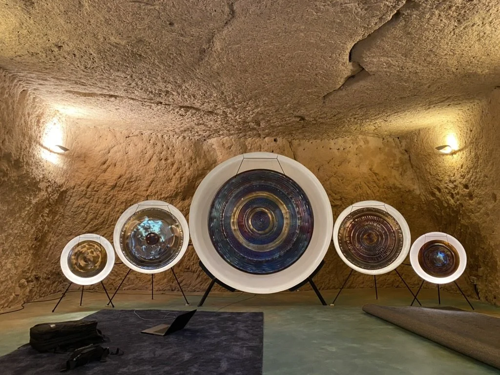 Five gongs of varying sizes mounted on stands in a cave-like space with stone walls and a carpet in front of the gongs for people to sit or lay down on.