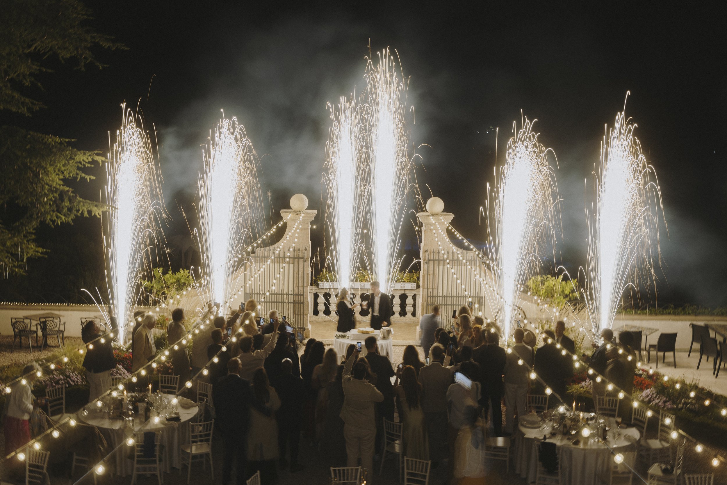 A nighttime wedding celebration with fireworks shooting up from a decorated backdrop with a historic wall and pillars in the background, surrounded by guests. Elegant tables are set up in the foreground, adorned with lights and floral arrangements.