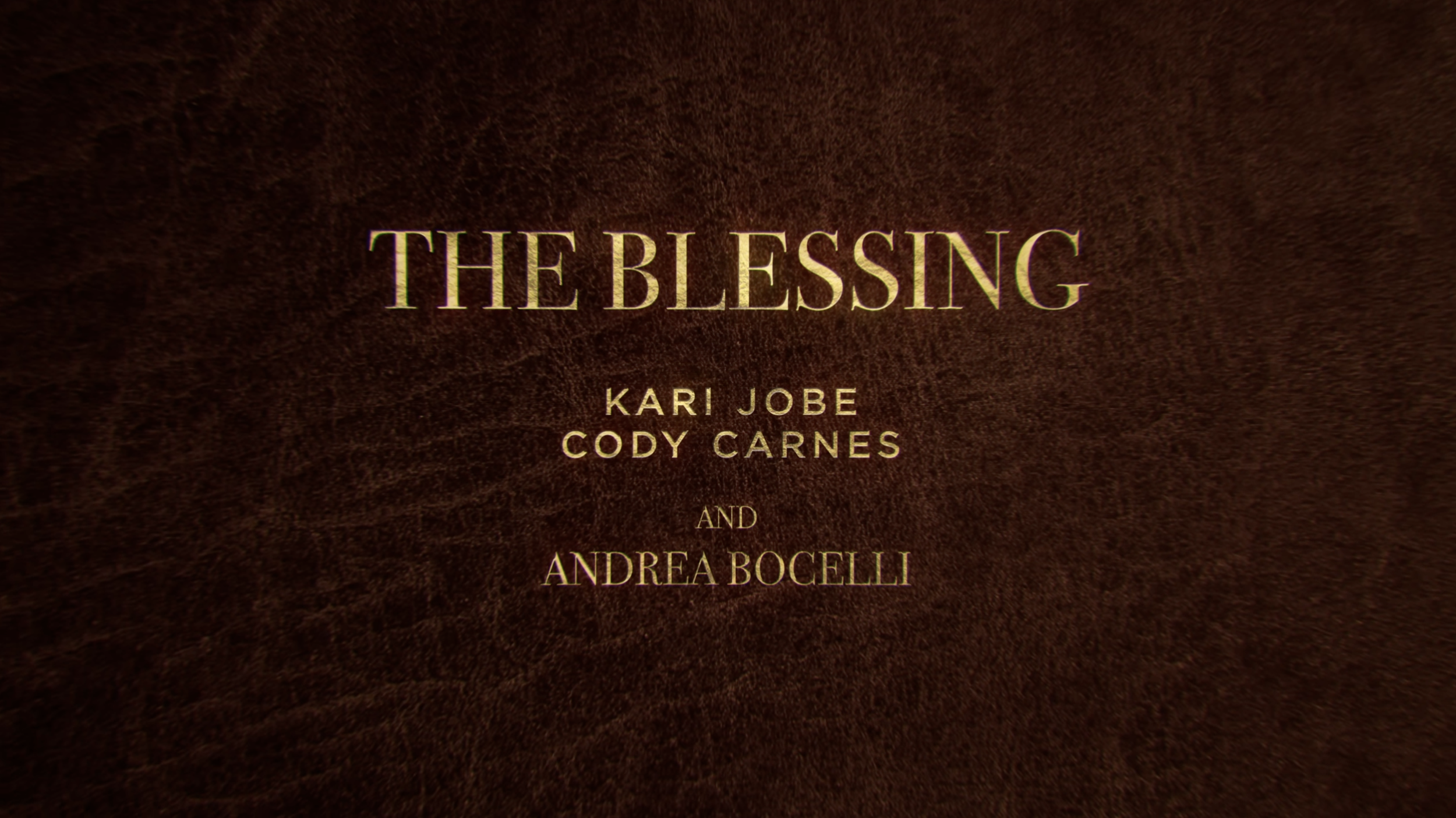 Screenshot of The Blessing lyric video by Kari Jobe and Cody Carnes, featuring Andrea Bocelli, with a brown leather background and golden lettering.