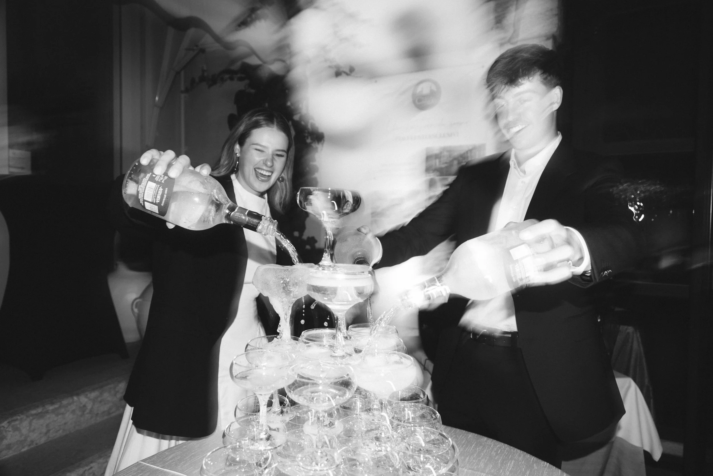 A black and white photo of a bride and groom pouring drinks into a tower of champagne glasses at a wedding, with a festive atmosphere in the background.