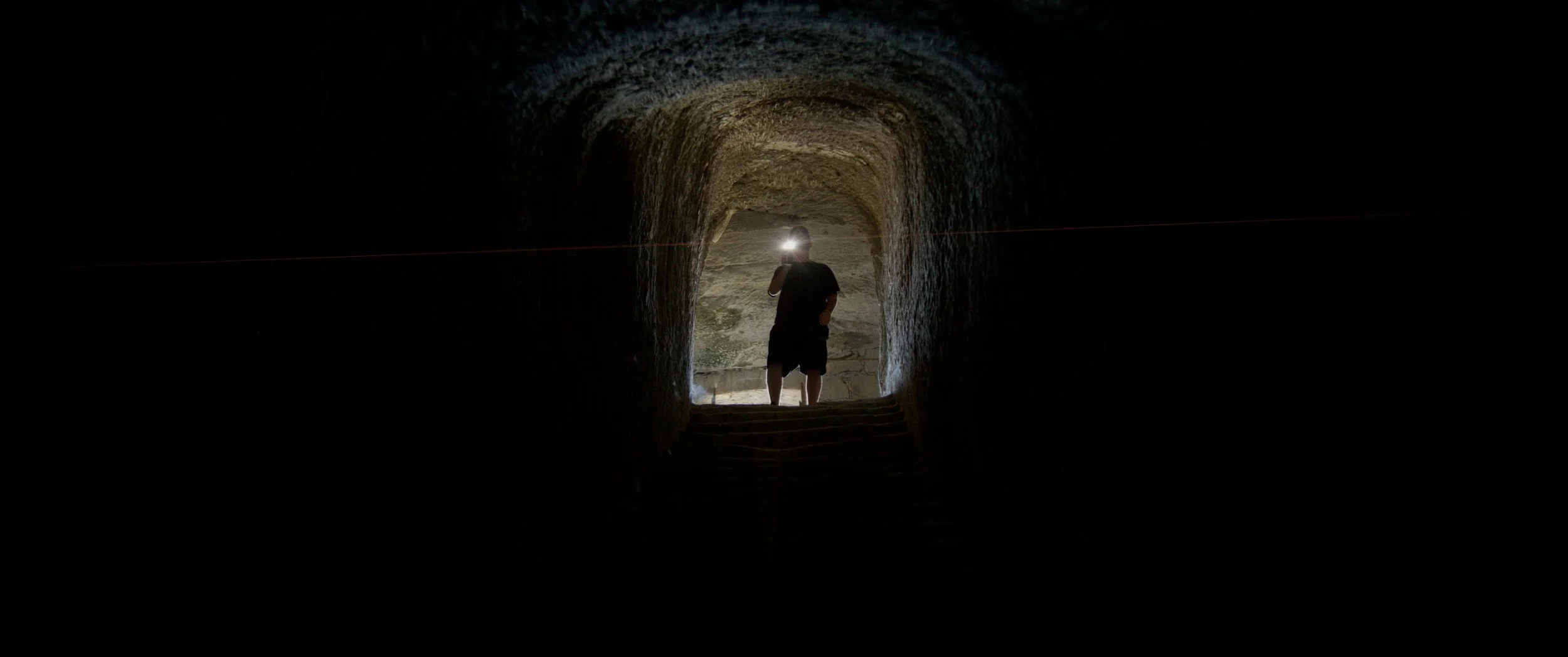 Person standing in a dark stone tunnel, facing towards the bright opening, with a head torch.