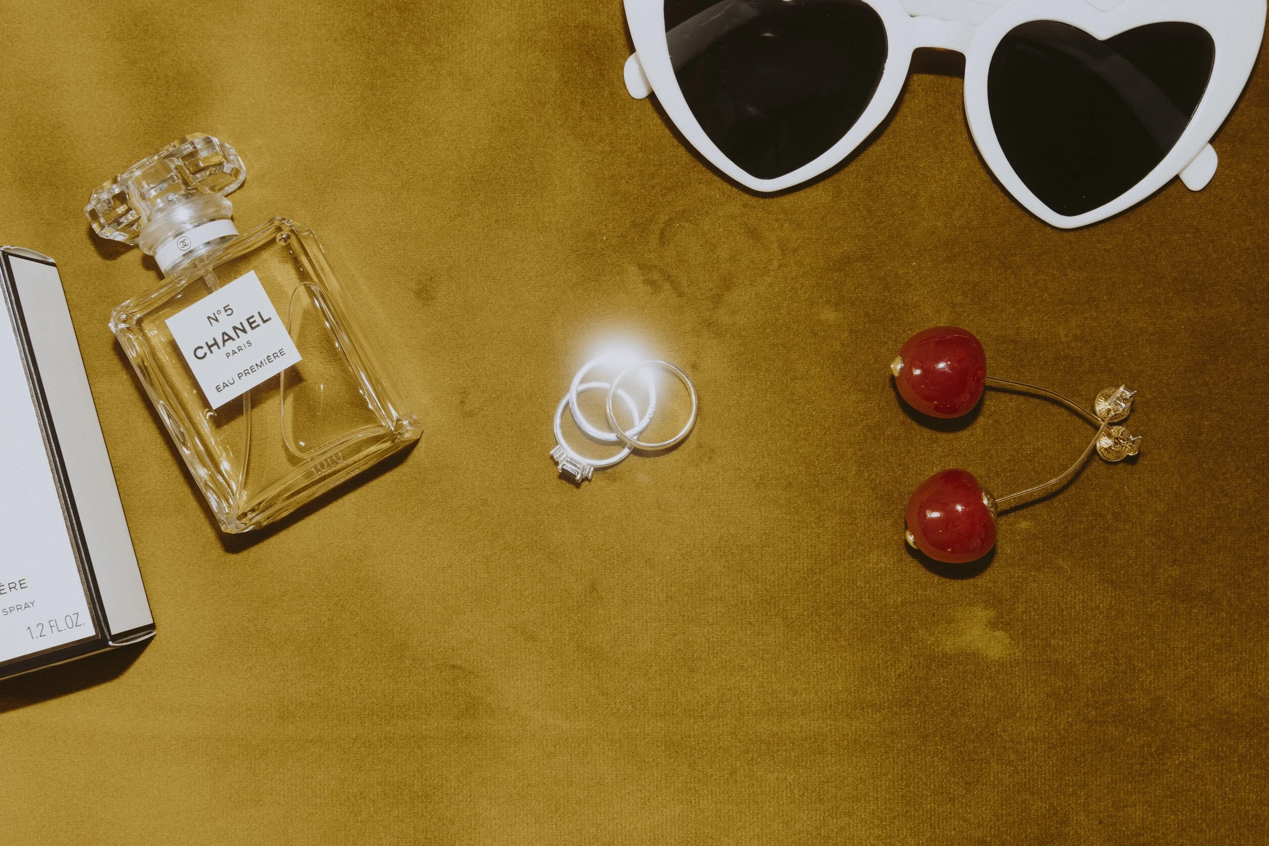 A flat lay of a Chanel perfume bottle, its box, silver wedding rings, red cherry-shaped golden and red earrings, and white heart-shaped sunglasses on a wooden surface.