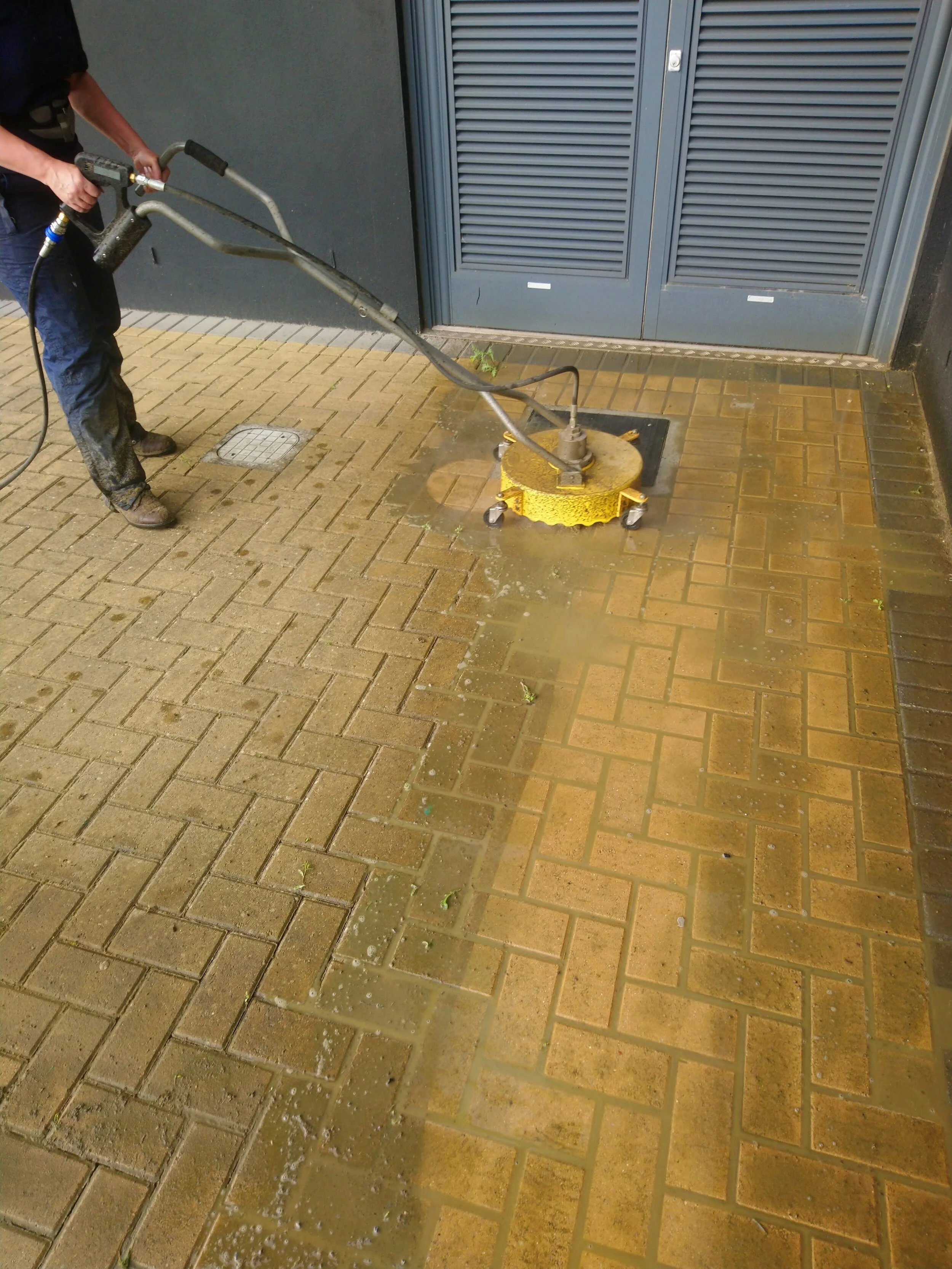 Person pressure washing a brown brick sidewalk near a blue metal door.