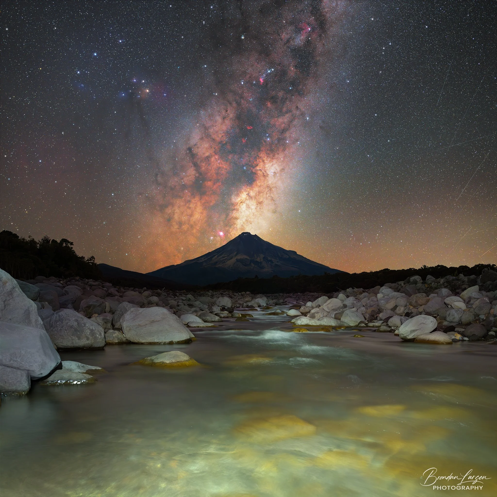 Nighttime scene with a starry sky, revealing the Milky Way galaxy above a mountain and a river with rocks.