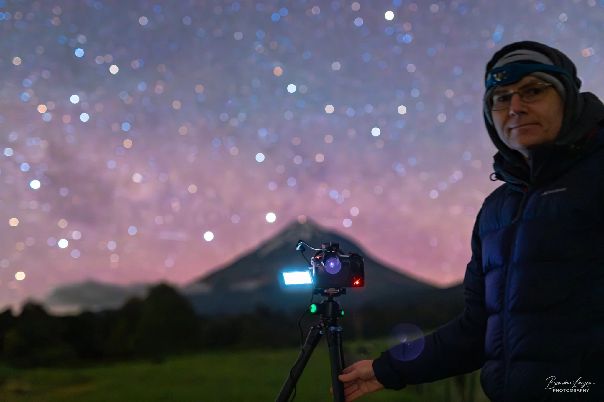 Photographer stands outdoors at night, with mountain and starry sky in background, holding a camera mounted on a tripod with a laptop and lights, capturing astrophotography.