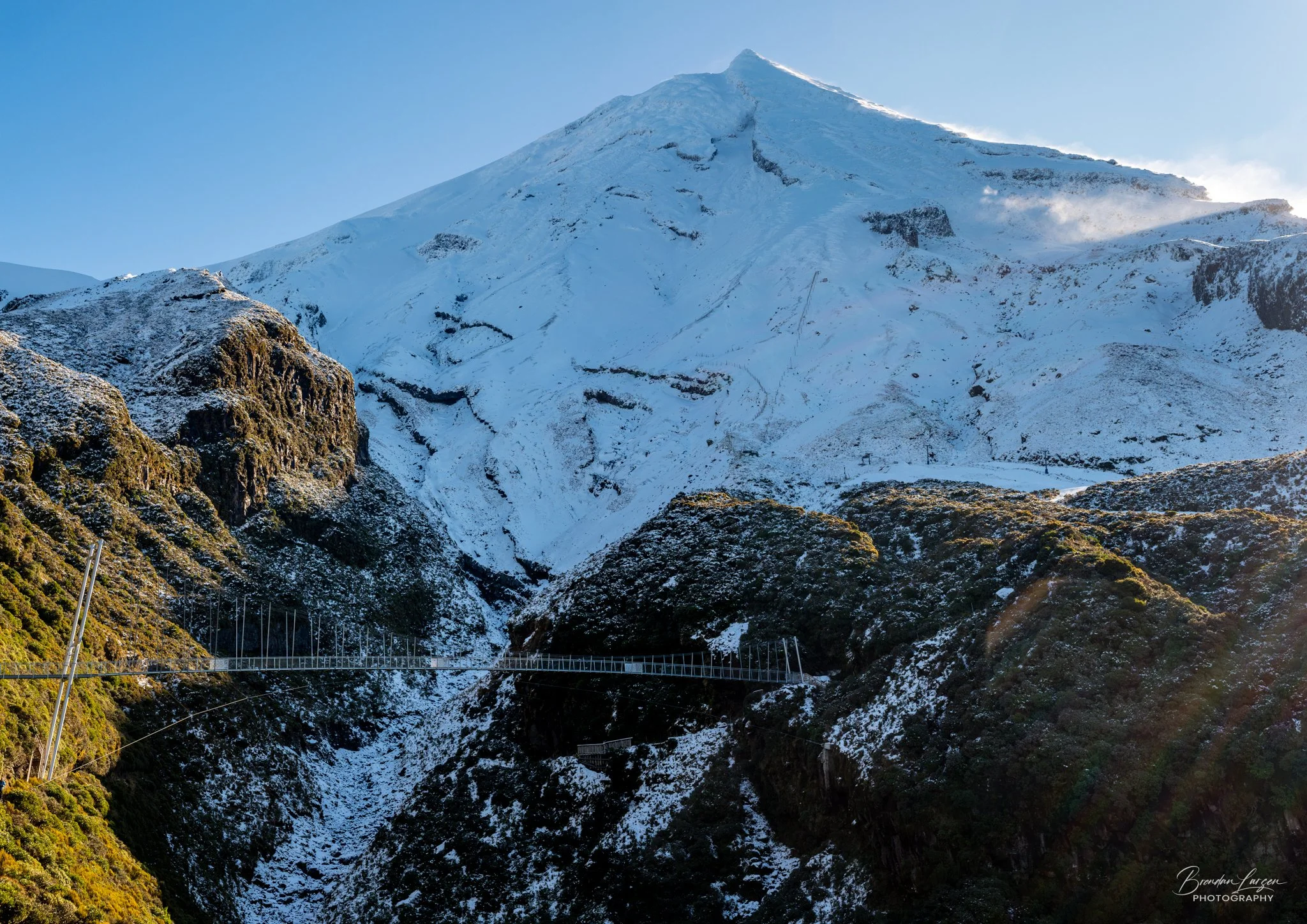 Snow-covered mountain with a suspension bridge in the foreground, lush green vegetation on the lower slopes, and blue sky.