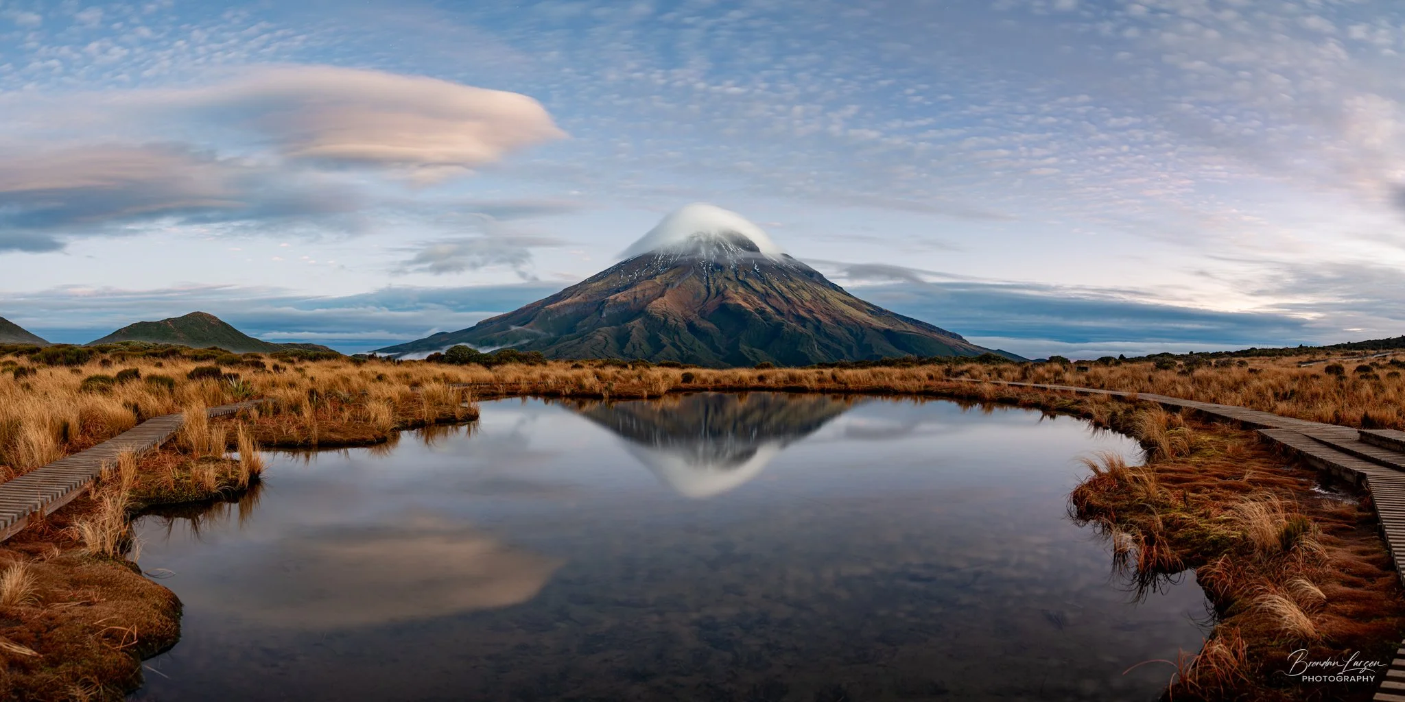 A mountain with a snow-capped peak reflecting in a calm pond in a grassy field under a partly cloudy sky.