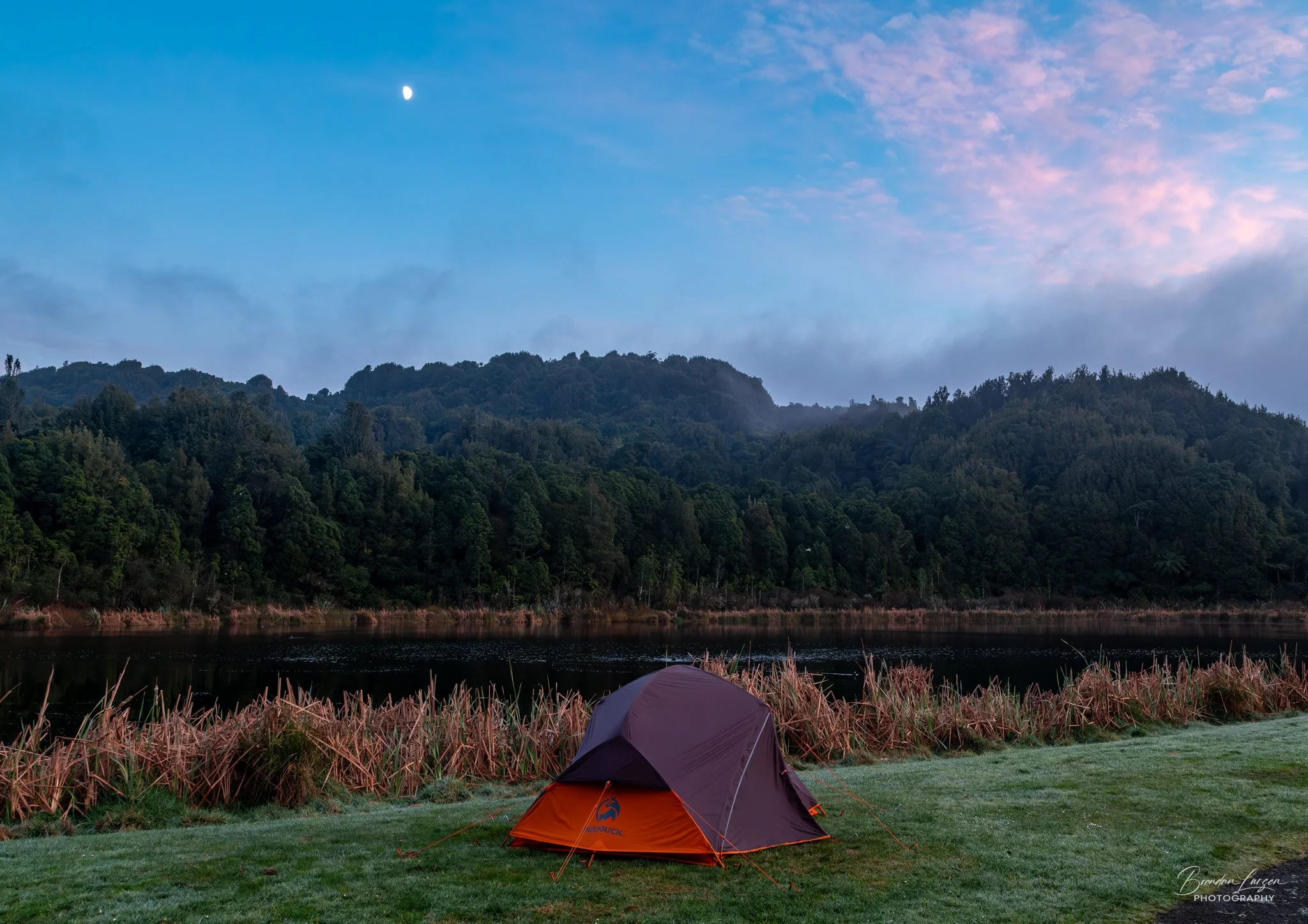 A tent pitched on a grassy area near a body of water, with a dense forest and hills in the background under a twilight sky with a visible moon.