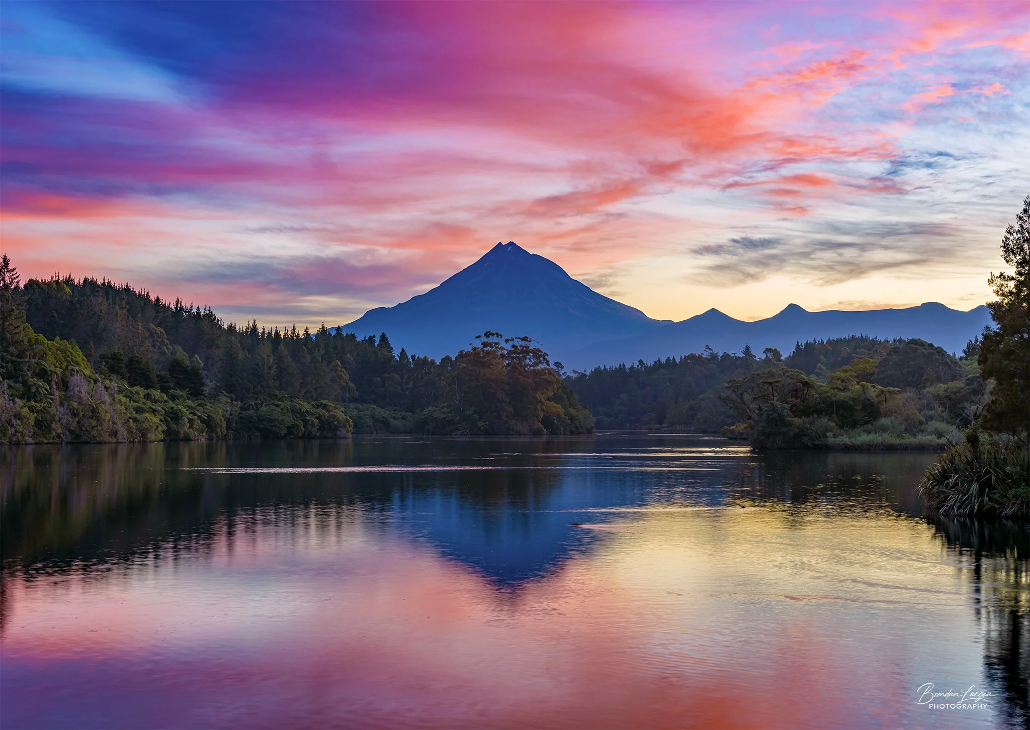 A scenic landscape of Mount Taranaki at sunset with a colorful sky, lush green trees, and a reflecting lake in the foreground.