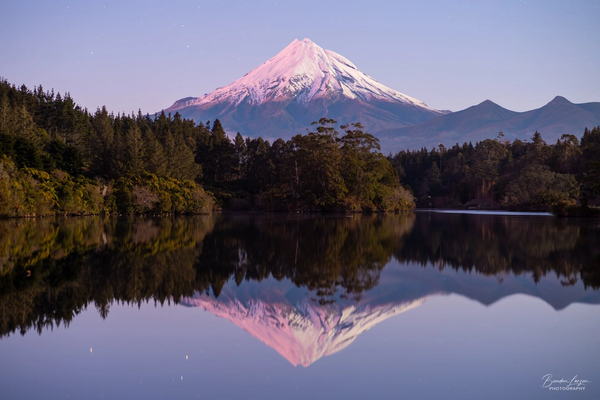 Snow-capped Mount Fuji reflected in a calm lake surrounded by dense forest under a clear purple sky.