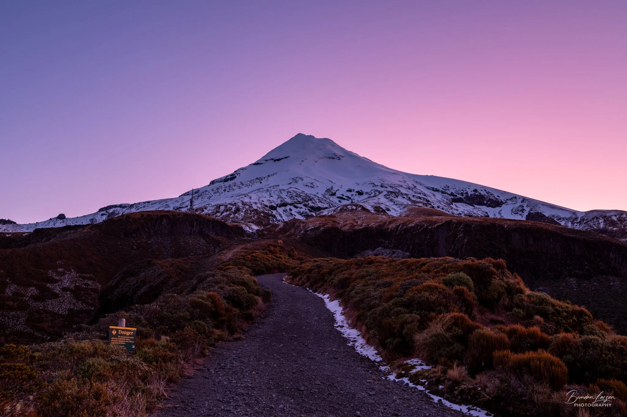 A mountain with snow at the top during sunset or sunrise, with a dirt trail leading up towards it, surrounded by bushes and a warning sign on the left side.