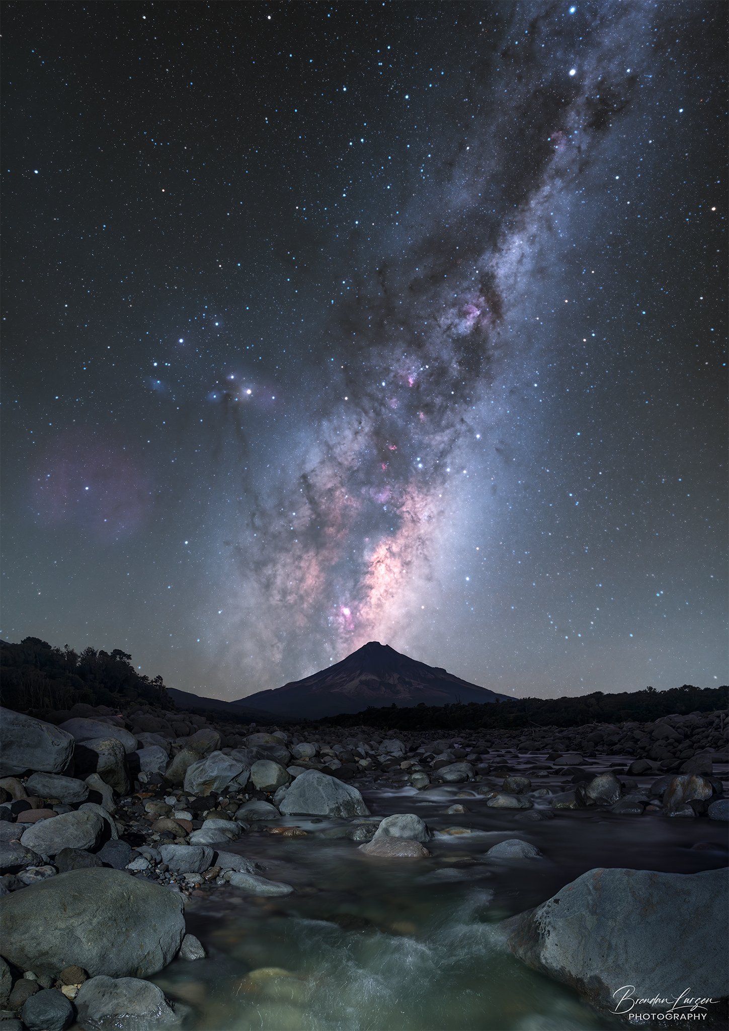 Milky Way galaxy visible in night sky over a mountain with a rocky river in the foreground.
