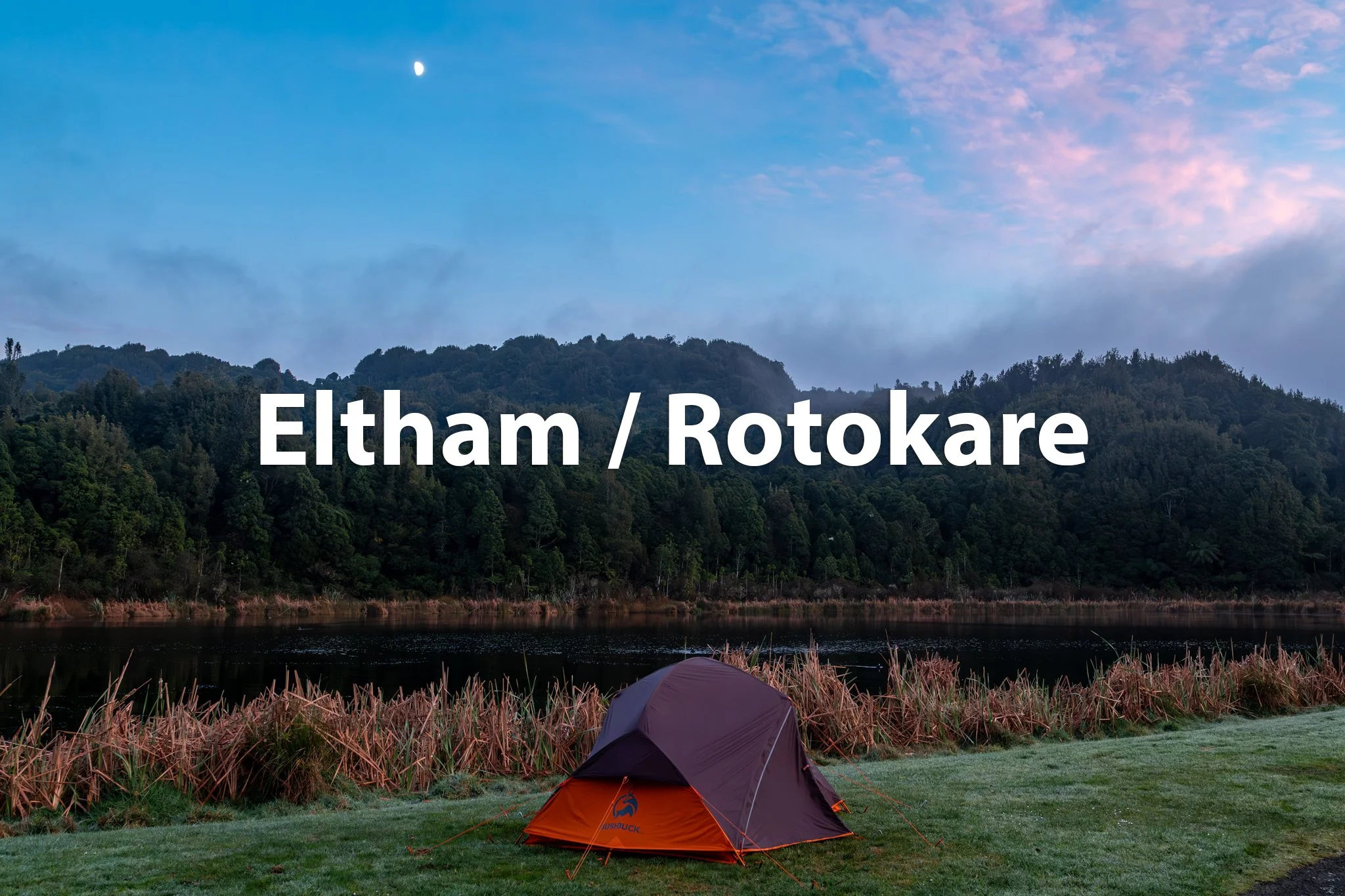 Tent pitched on grassy area near a body of water with forested hills in the background, a darkening sky with a moon.