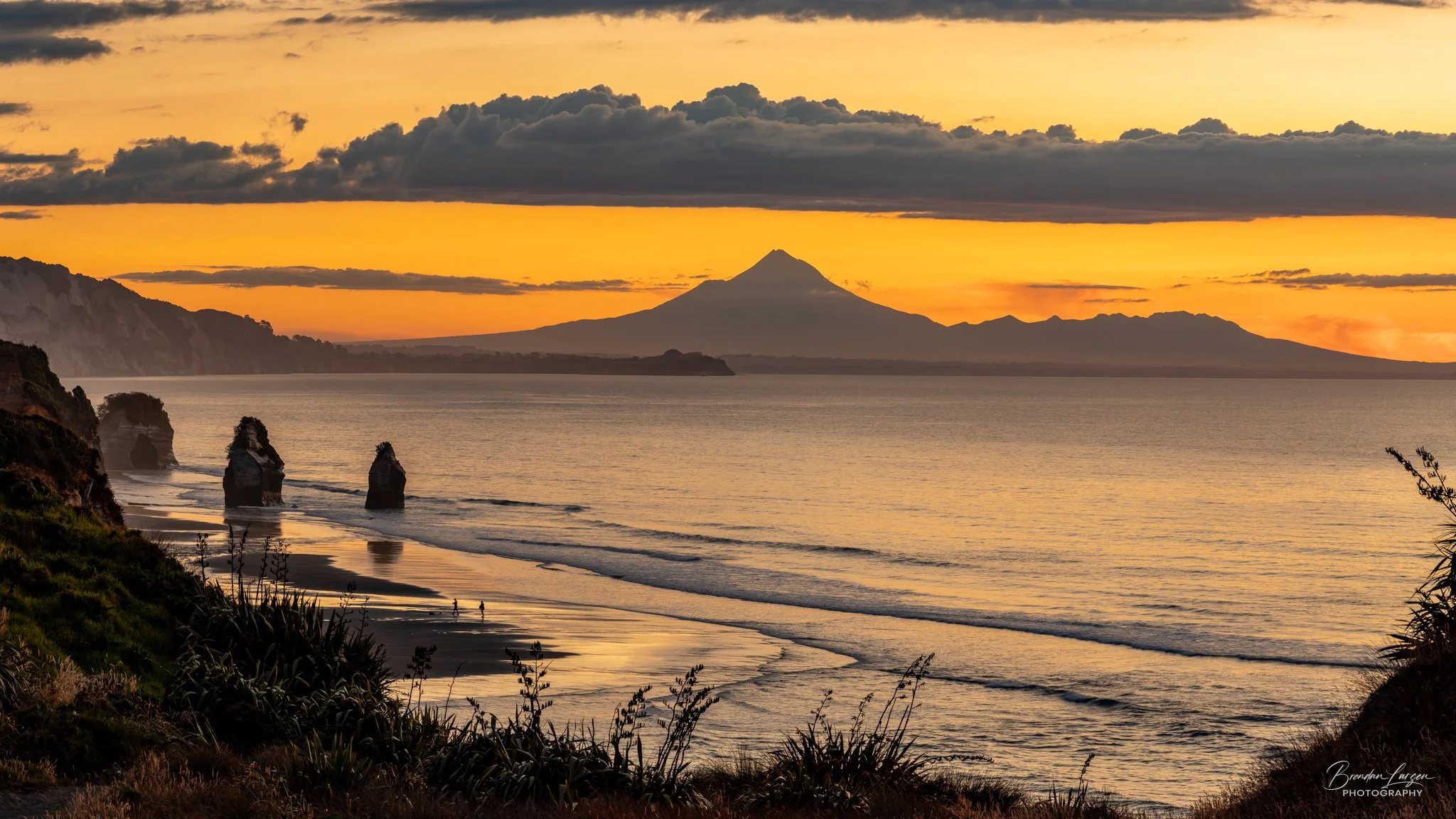 Sunset over the ocean with a volcanic mountain in the distance, three sea stacks along the shoreline, and clouds in the sky.
