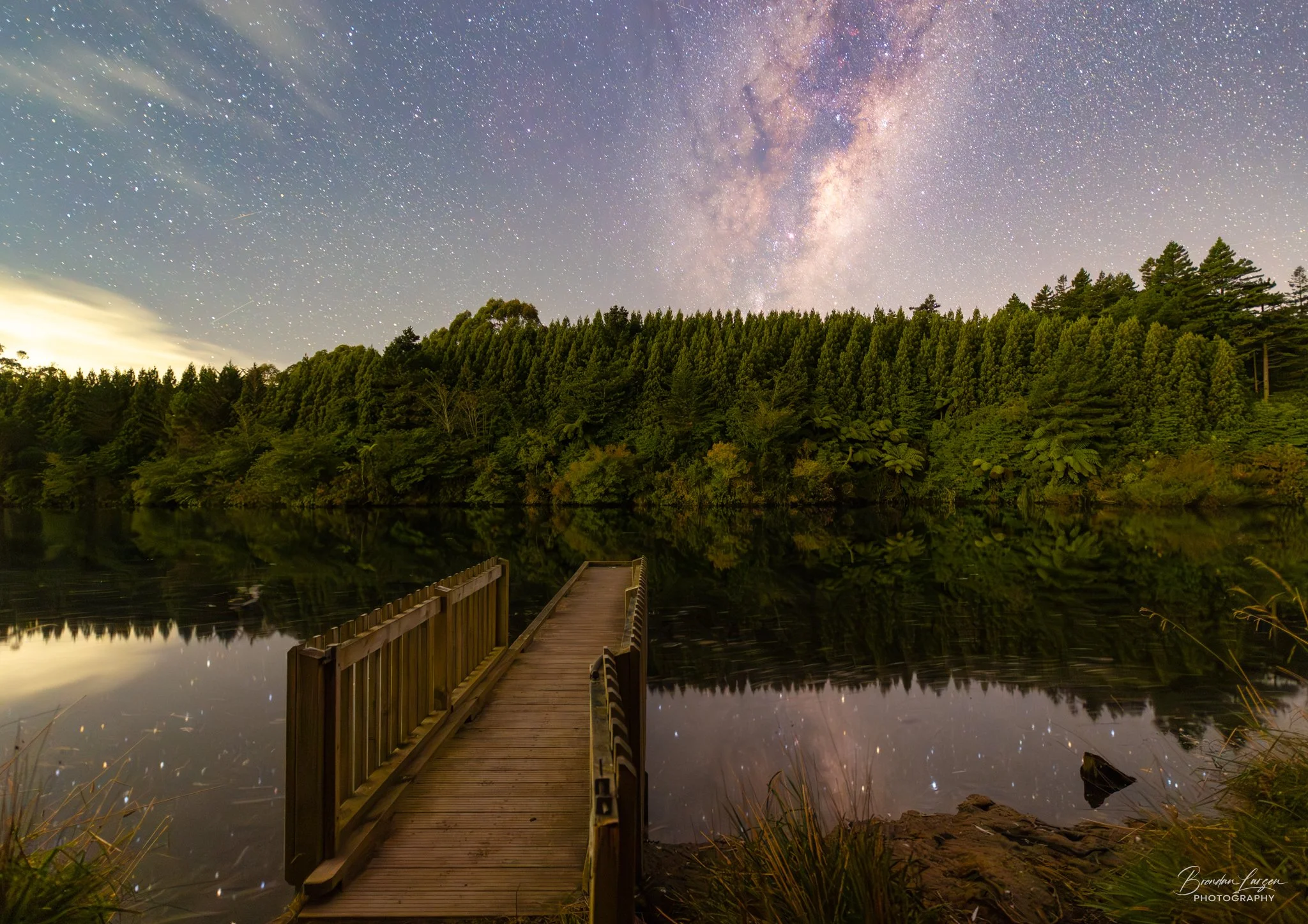 A wooden dock extends into a calm body of water surrounded by lush green trees, with a starry night sky and the Milky Way galaxy visible overhead.