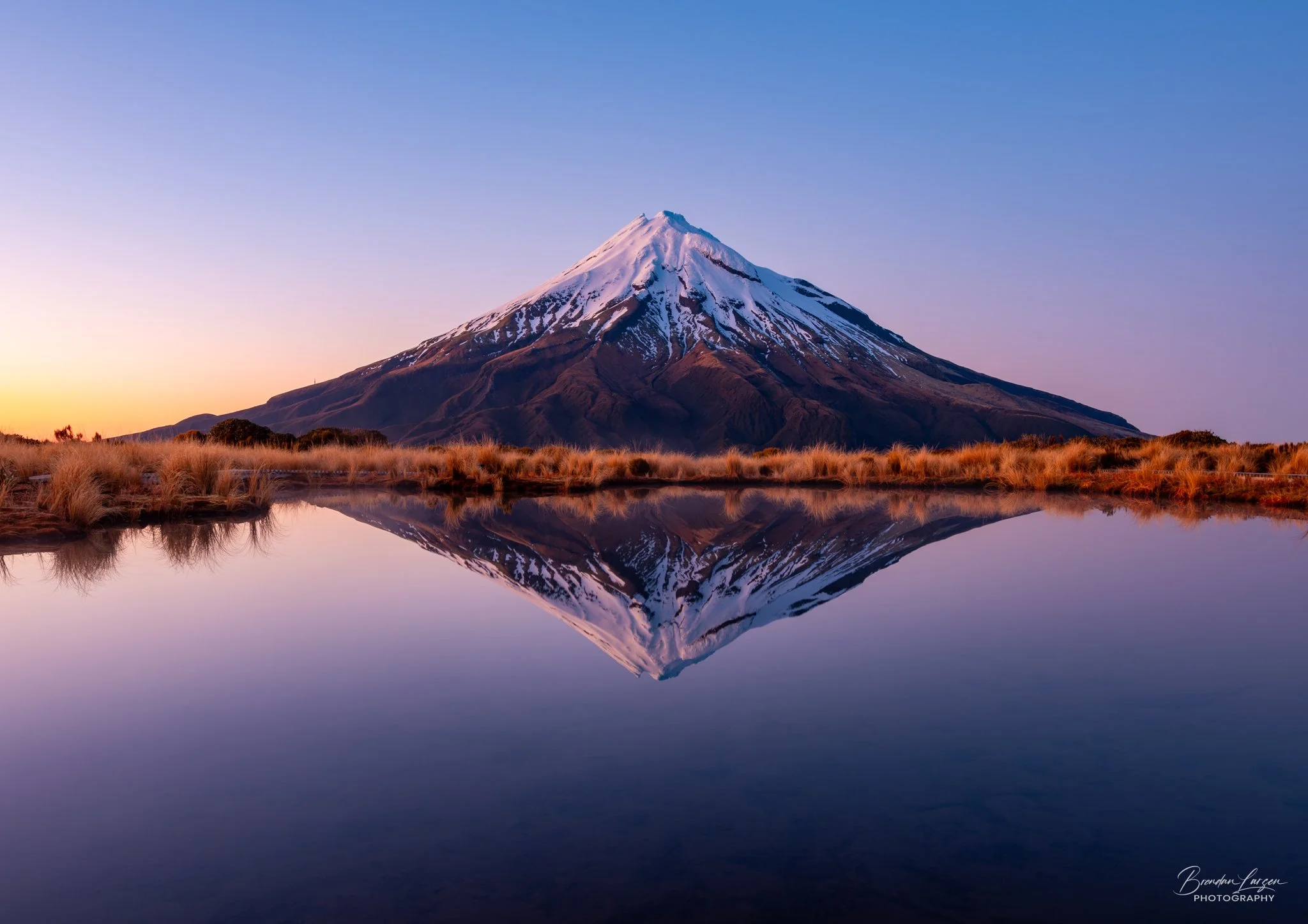 Snow-capped mountain reflected in calm water with grassy shoreline at sunrise or sunset.