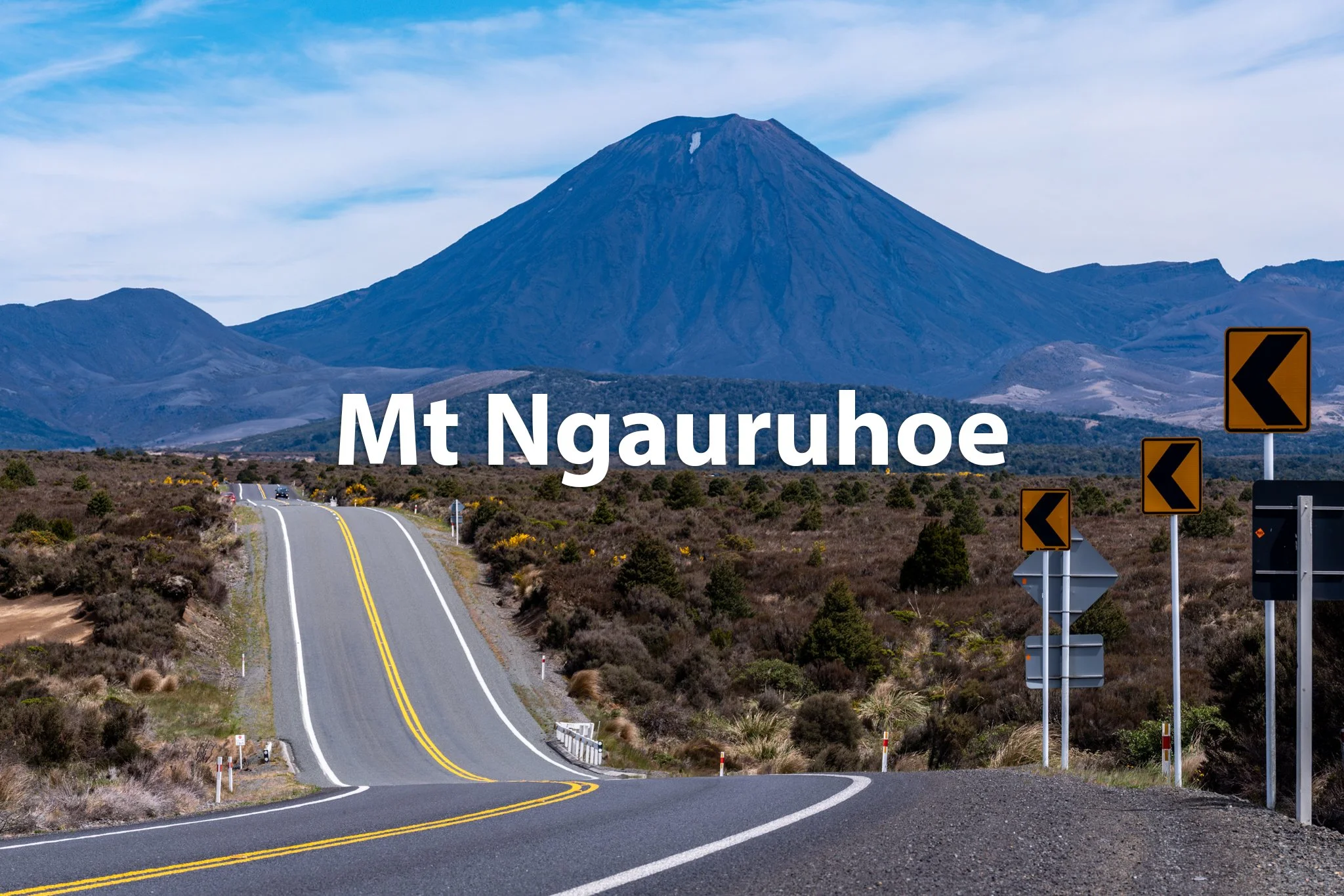 Scenic view of Mt. Ngaruhoe with a winding road in the foreground, yellow warning signs, and cloudy sky.