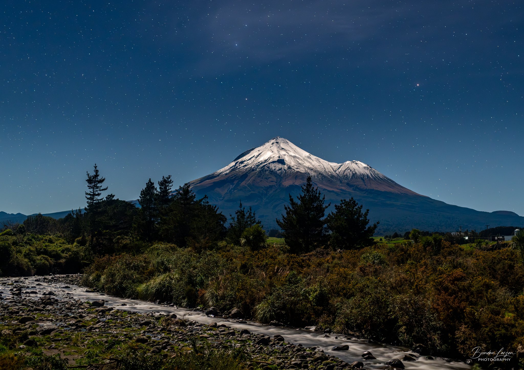 Night landscape featuring a snow-capped mountain under a starry sky, with trees and a river in the foreground.