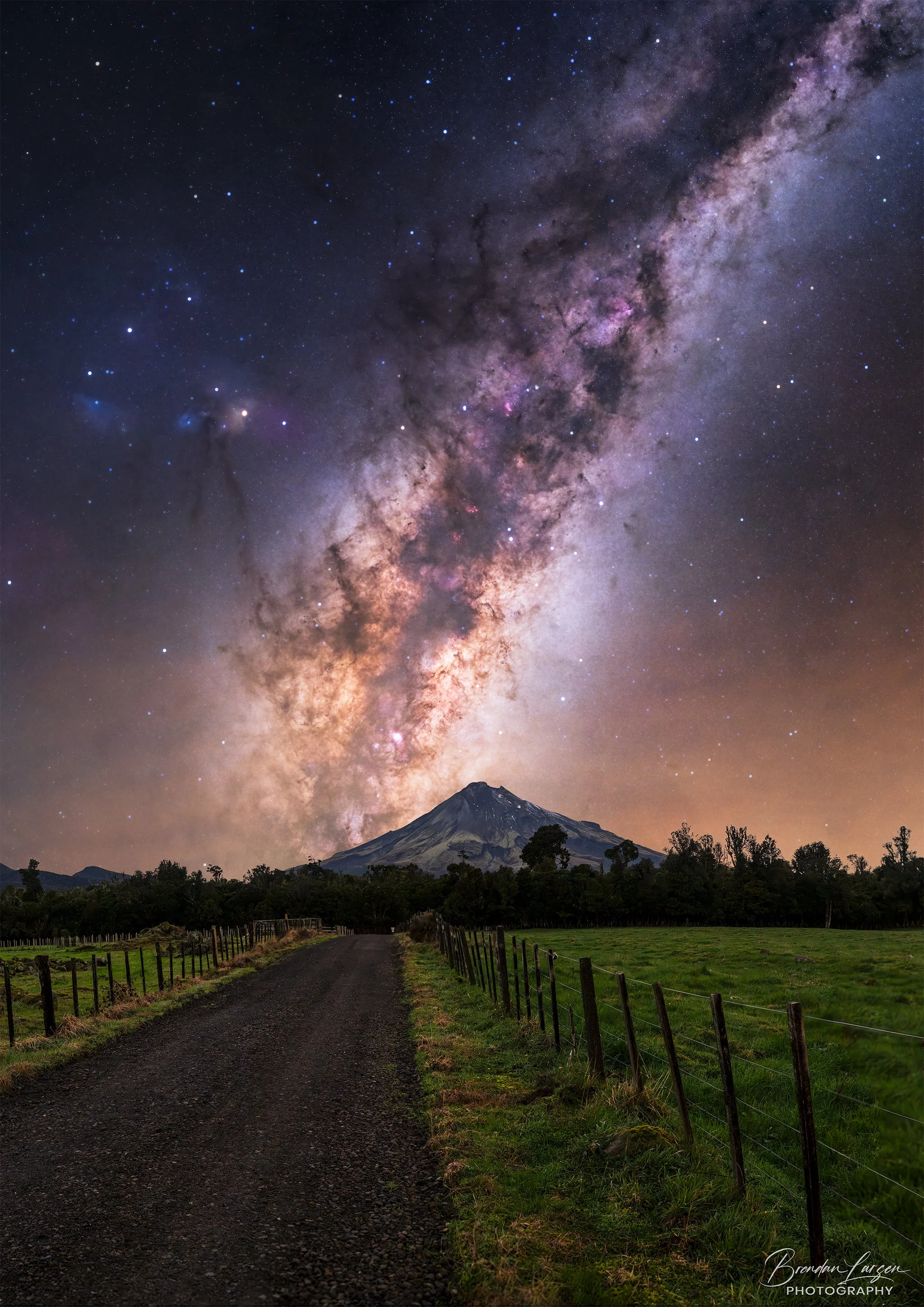 Night sky showing the Milky Way galaxy over a mountain, with a dirt road leading toward the mountain and fenced grassy fields on either side.